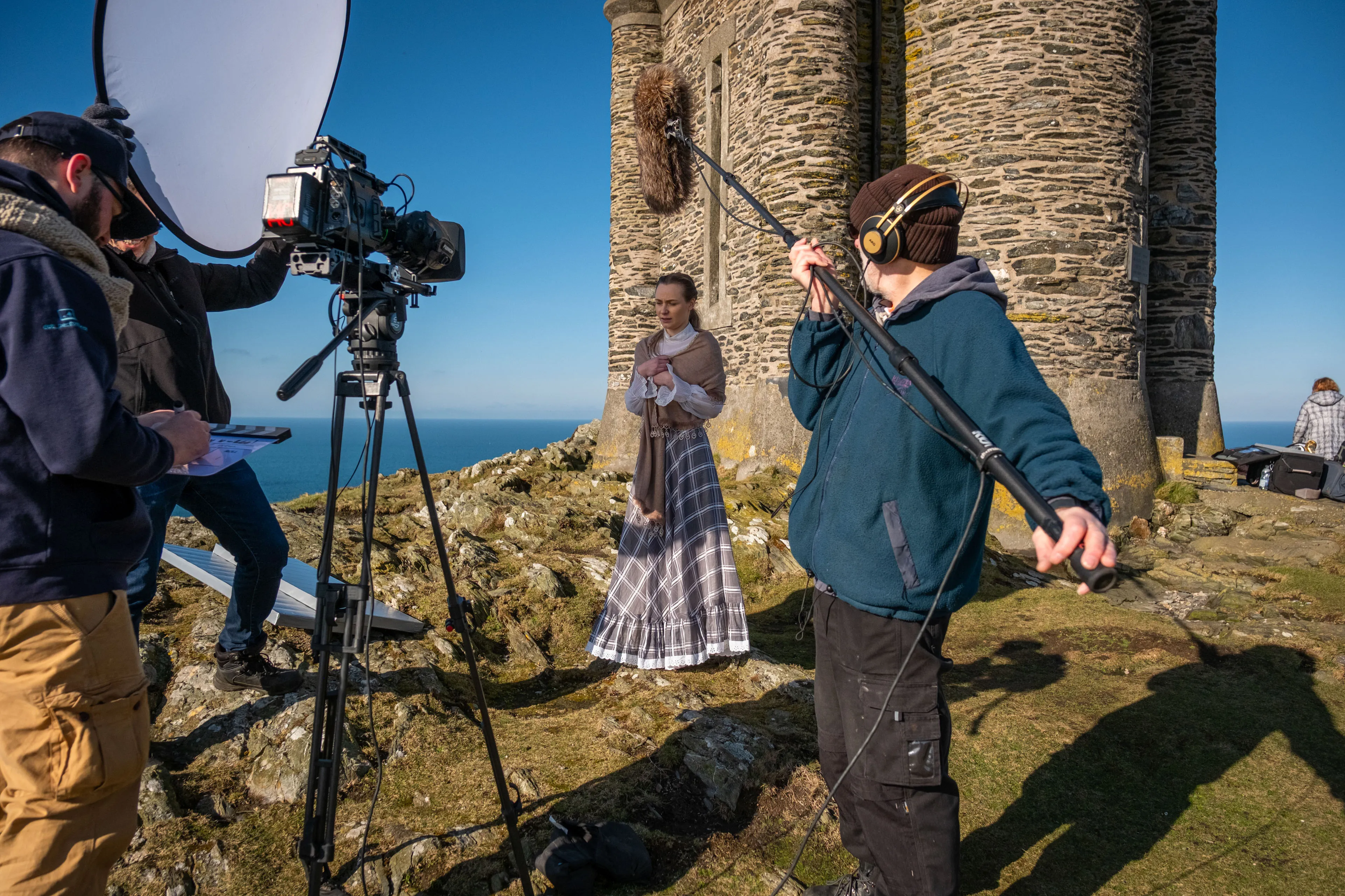 Nico van Loggerenberg, Dave Armstrong, Alice Smith and Richard Fryer filming on location Milners tower on the Isle of Man coastline, with an URSA Mini Pro 12K camera on tripod, reflector dish and boom operator visible alongside a performer in period costume