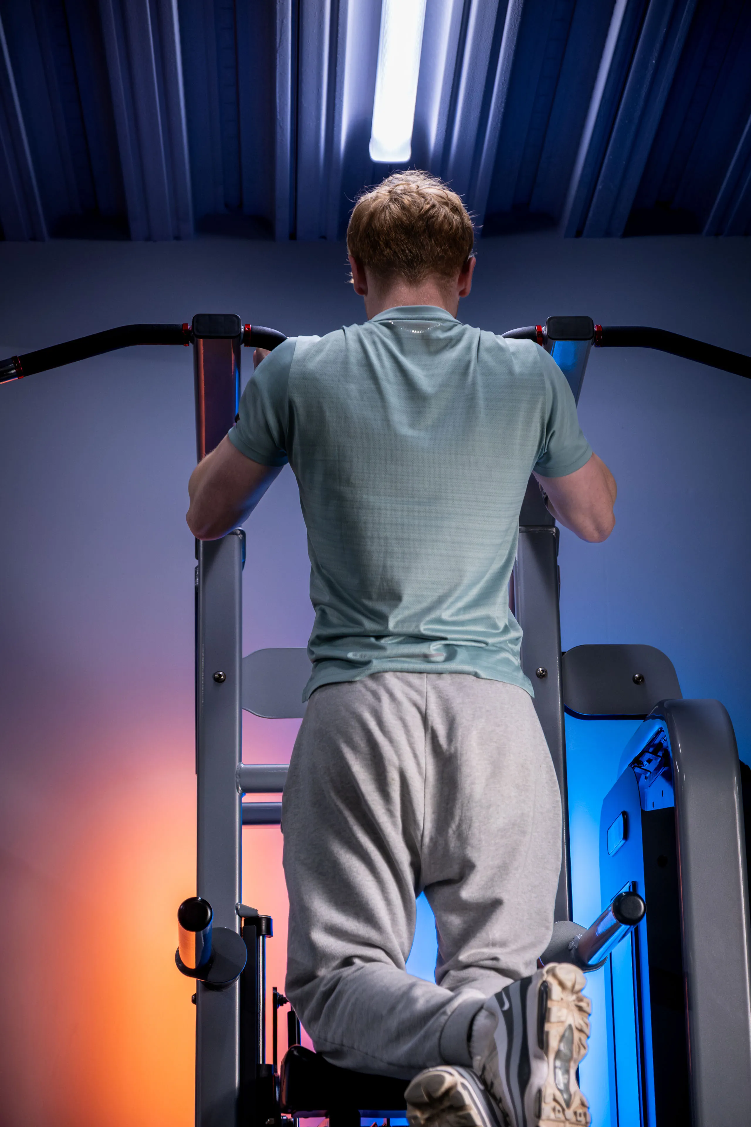Man doing pull-ups in a gym, wearing a blue shirt and gray sweatpants. The room is lit with blue and orange lights, creating a focused atmosphere.