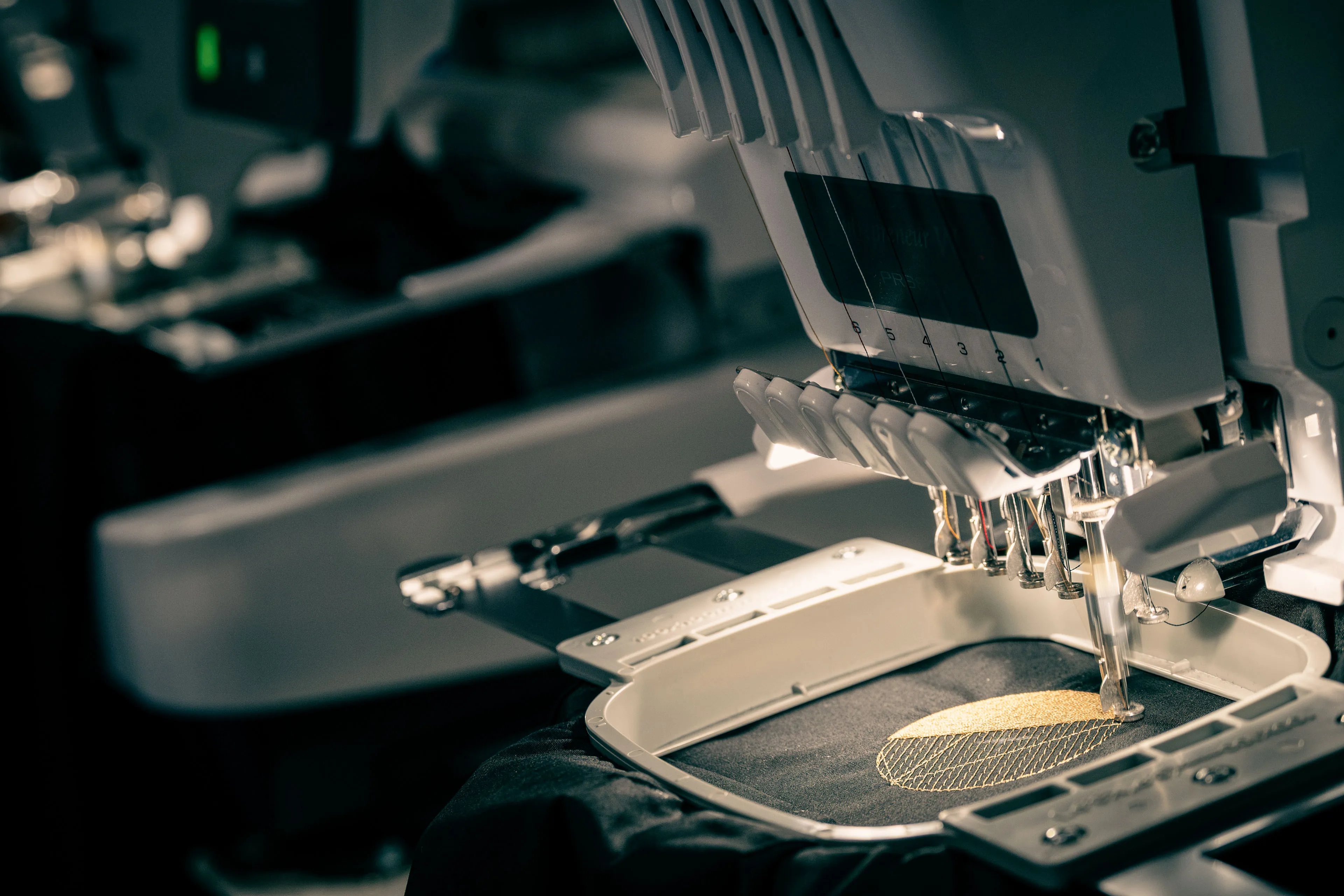 Close-up of an embroidery machine in dim lighting, focused on a gold thread design being stitched on black fabric, conveying precision and craftsmanship.