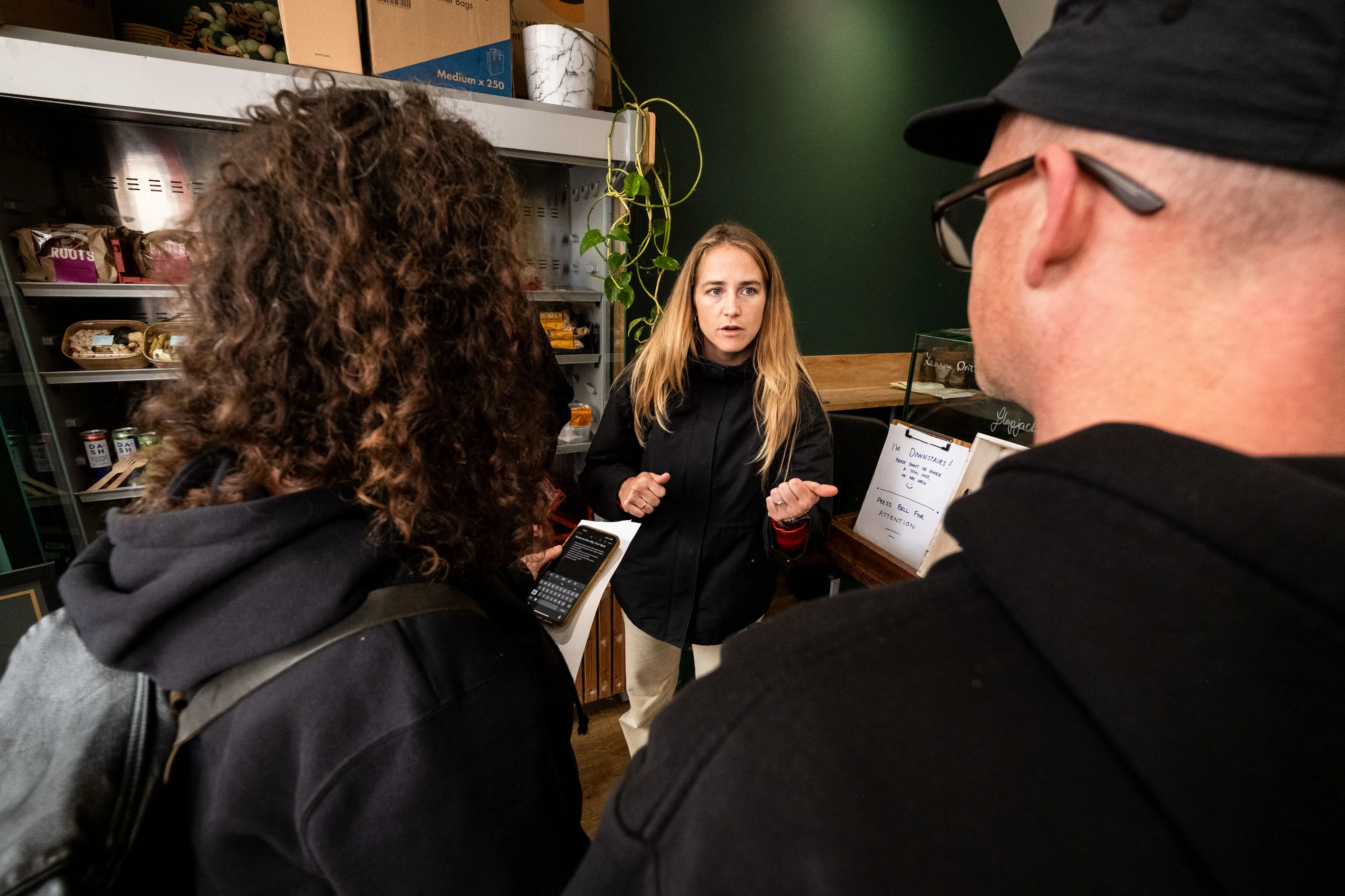 A woman with long blonde hair speaks assertively to two people in a store. Shelves with products are visible in the background, creating a busy, focused atmosphere.