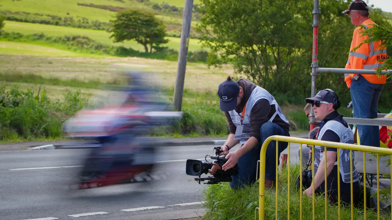Will Oates holds an URSA 12K and films as TT riders speed past. Juan Readshaw is recording sound. Both wear white TT approved tabards that allow for closer access to the races.