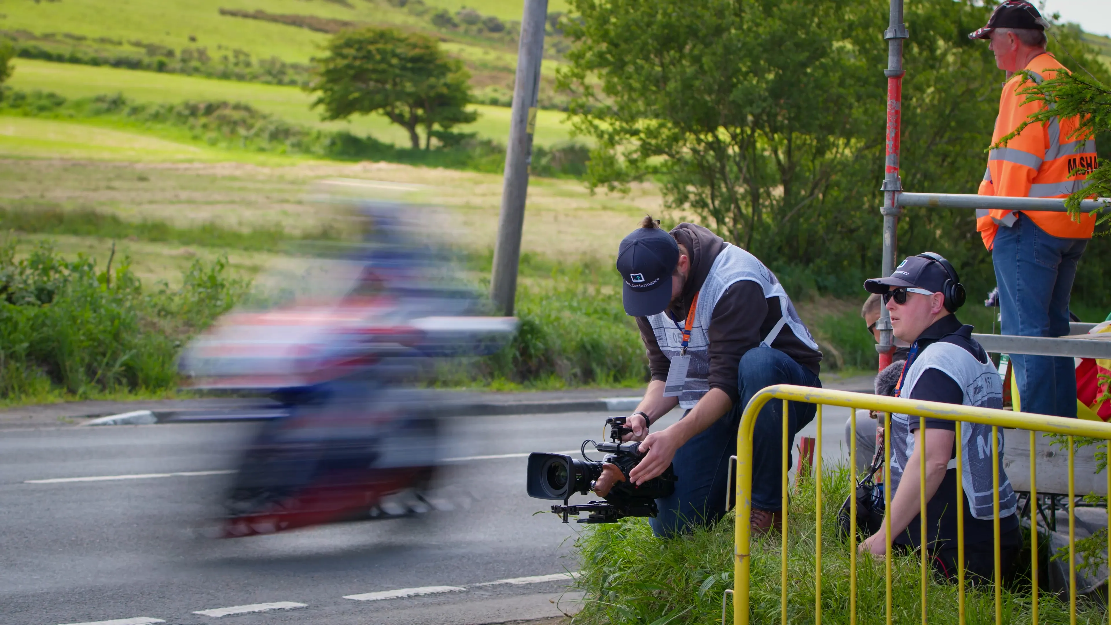 Will Oates holds an URSA 12K and films as TT riders speed past. Juan Readshaw is recording sound. Both wear white TT approved tabards that allow for closer access to the races.