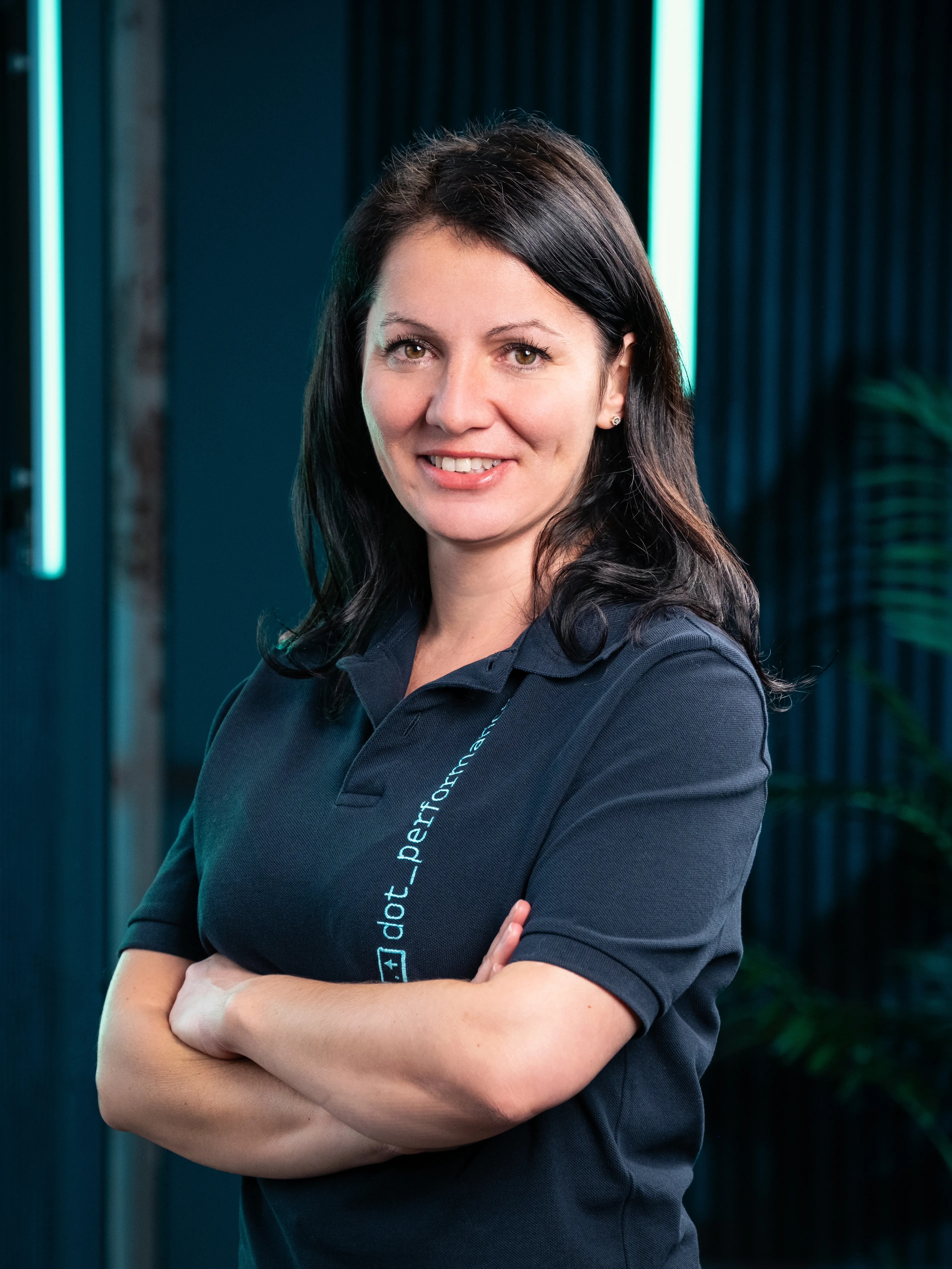 Headshot of Dot Performance's Operations Director Raluca Matei. She is wearing a navy branded polo shirt and is set against a dark wall with blue neon lights.