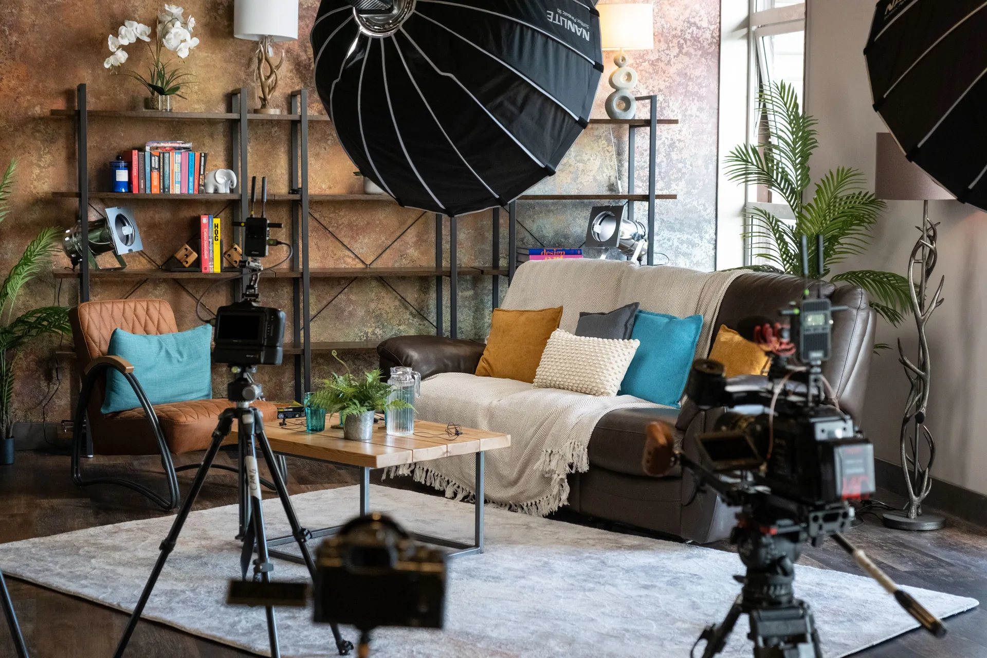 Full DotPerformance podcast and interview studio set on the Isle of Man showing multiple cinema cameras on tripods, Nanlite softbox and umbrella lighting, a sofa and seating area with cushions, and an industrial shelving backdrop