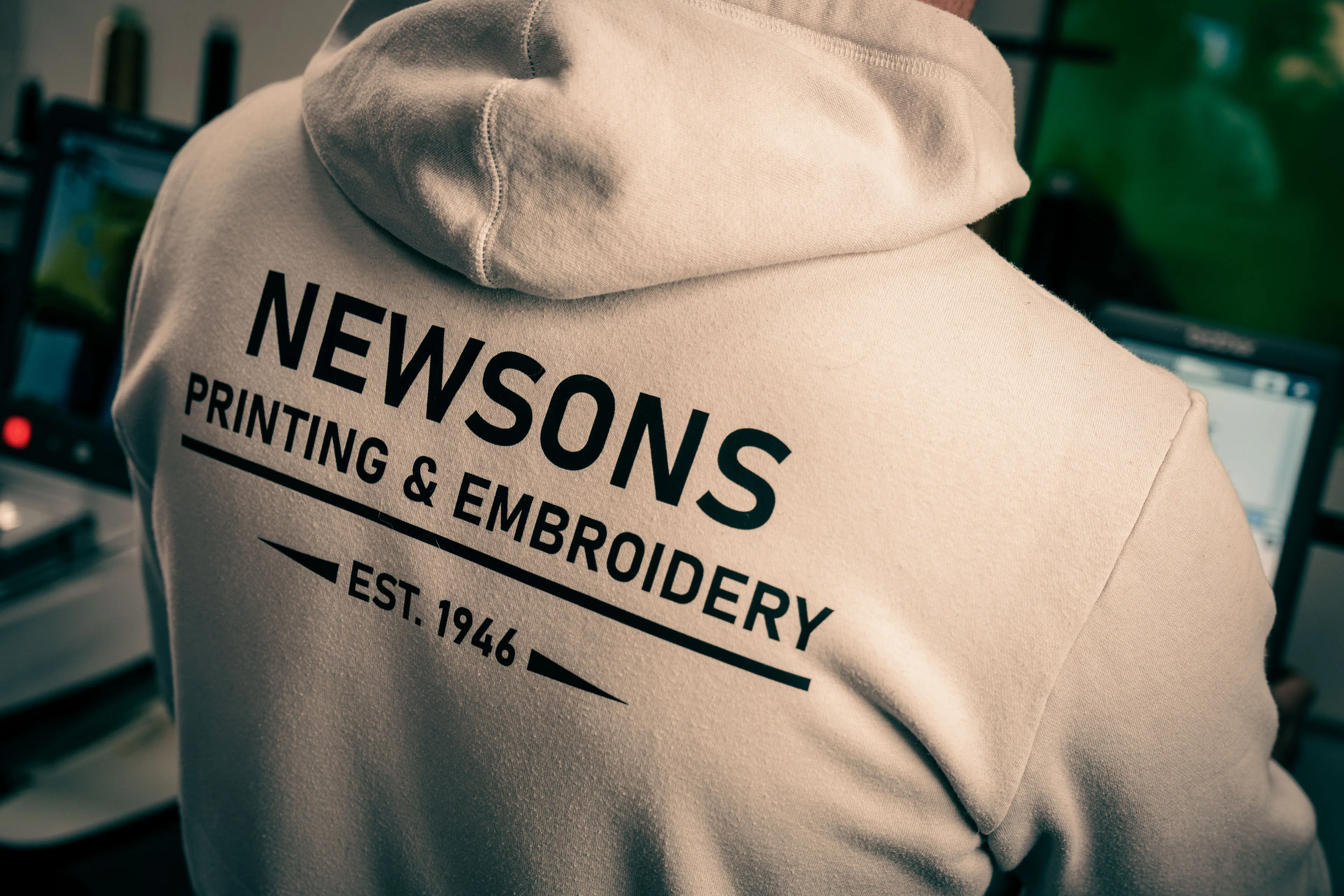A person wearing a hoodie with "Newsons Printing & Embroidery, Est. 1946" printed on the back. The setting appears to be an office with computer screens.