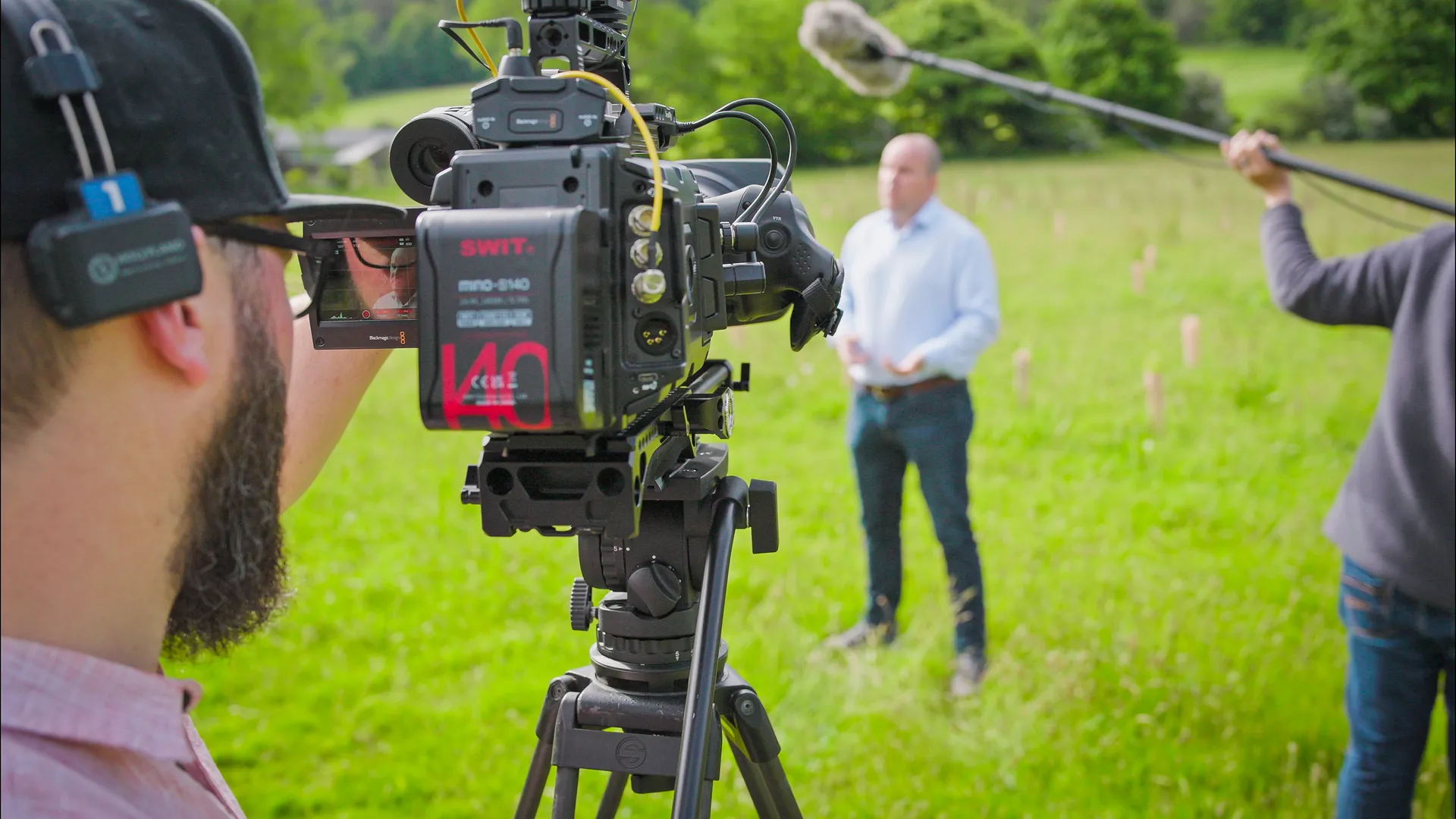 A cameraman films a man speaking outdoors in a lush green field. Another person holds a boom microphone. The setting conveys a professional video shoot.