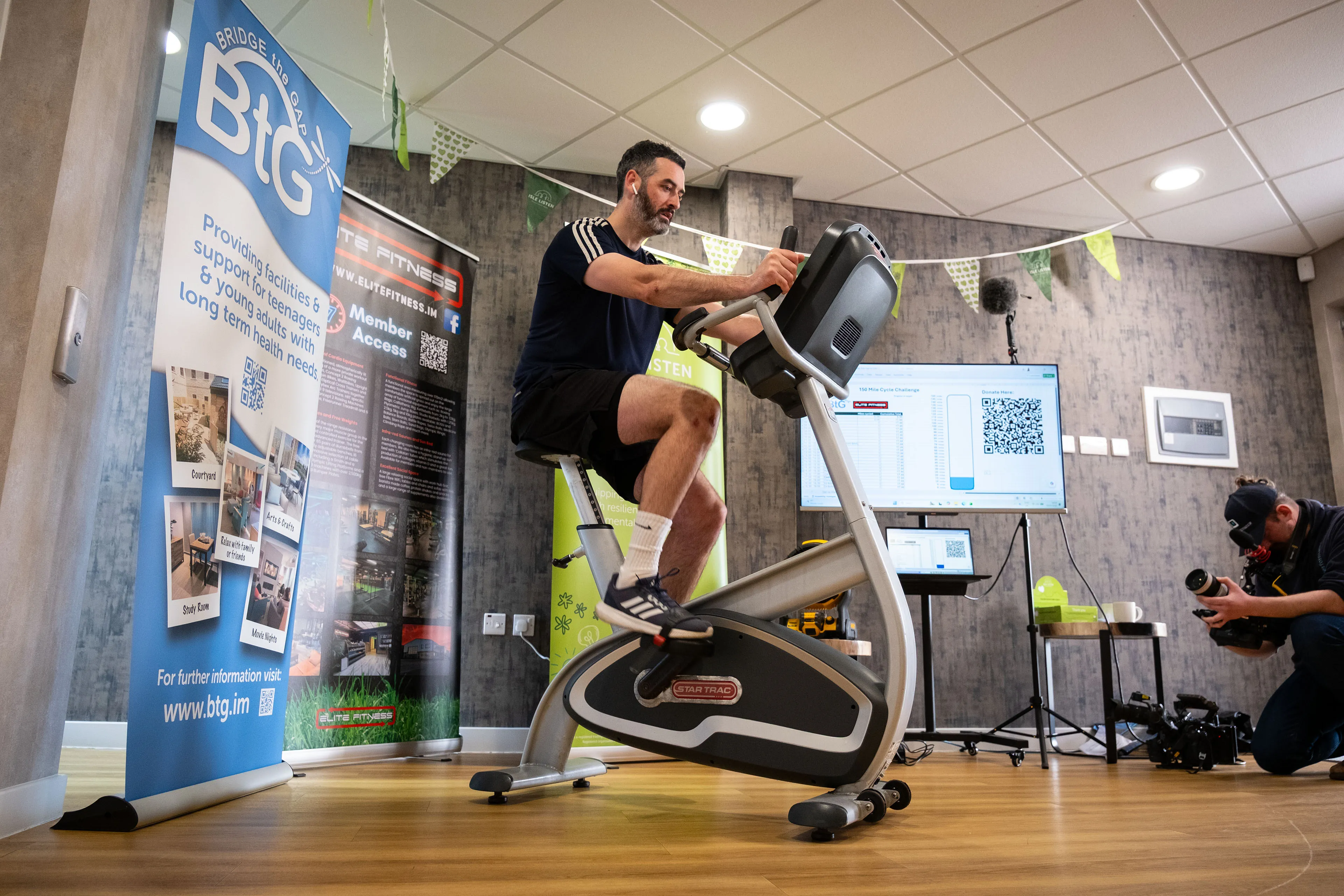 Dean Kearns cycling on a static bike during the 12-hour charity challenge while Will Oates from DotPerformance films in the background with a Black Magic pocket 6K