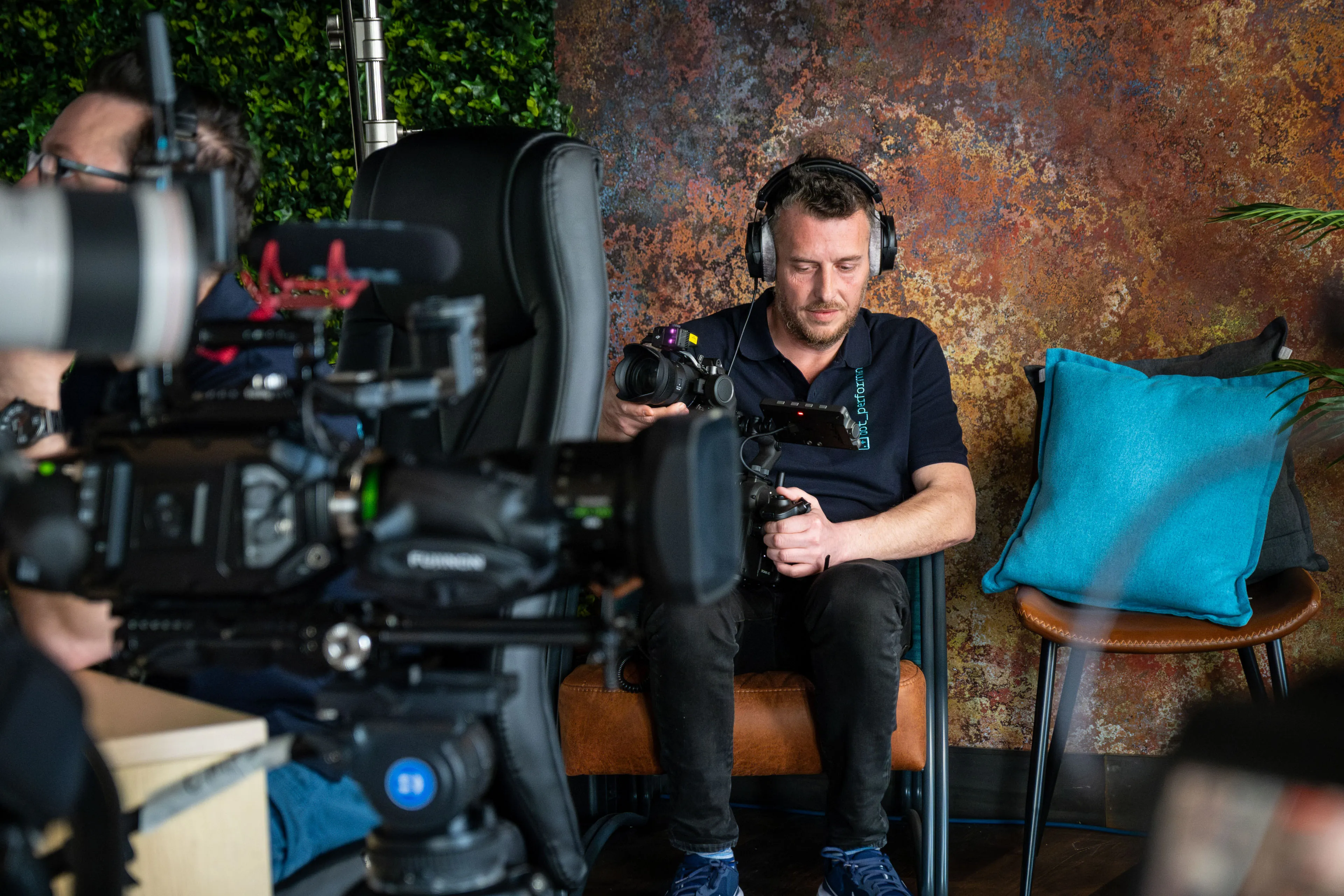 Andy Orton wearing headphones checking the DJI Ronin 4D camera monitor while seated in the studio during a multi-camera podcast production on the Isle of Man, with additional cameras visible in the foreground
