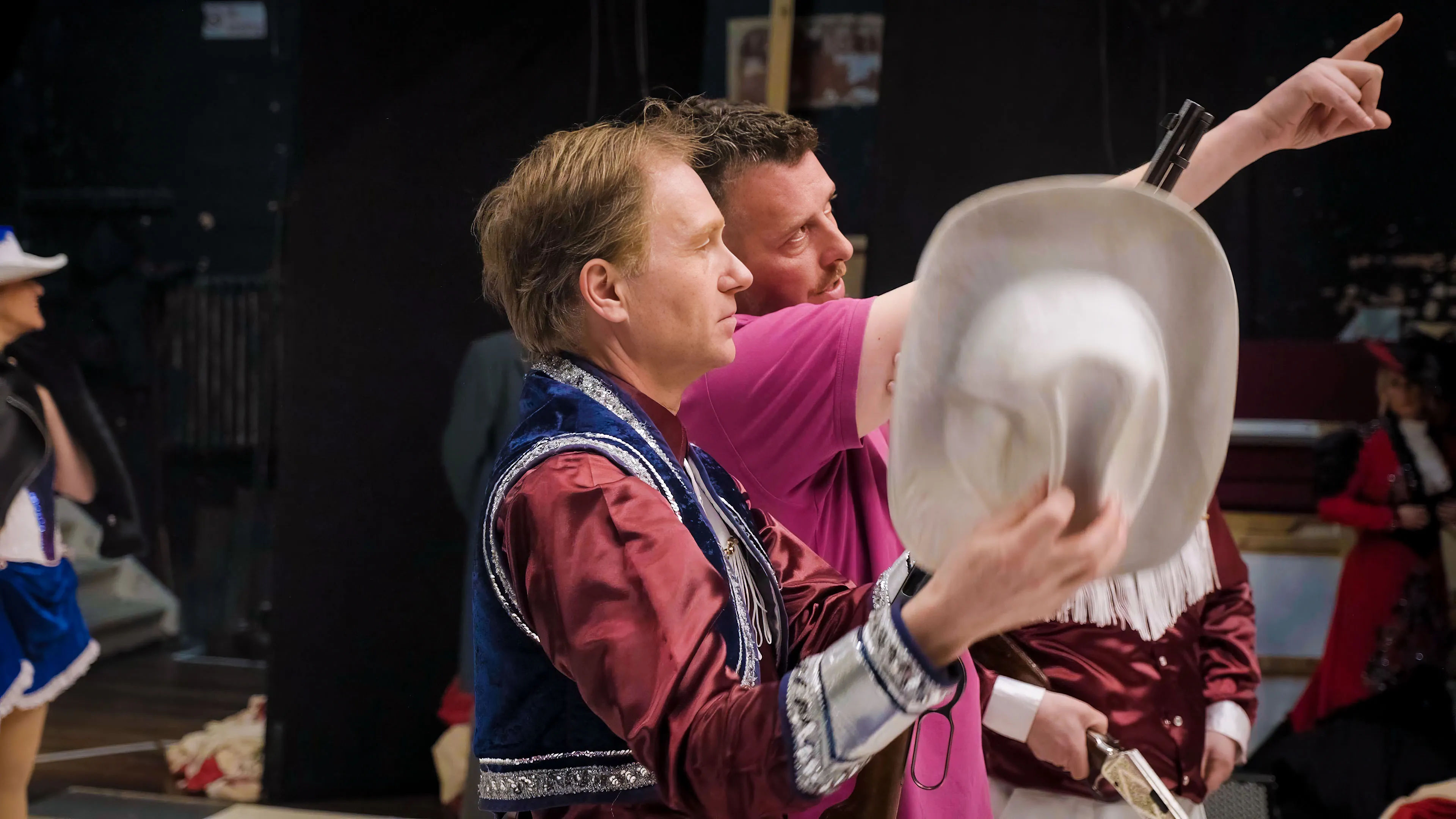 Two men are backstage at a theater. One wears a shiny costume with blue and silver details, holding a white cowboy hat. The other, in a pink shirt, points ahead, discussing something intently. The scene conveys preparation and focus.