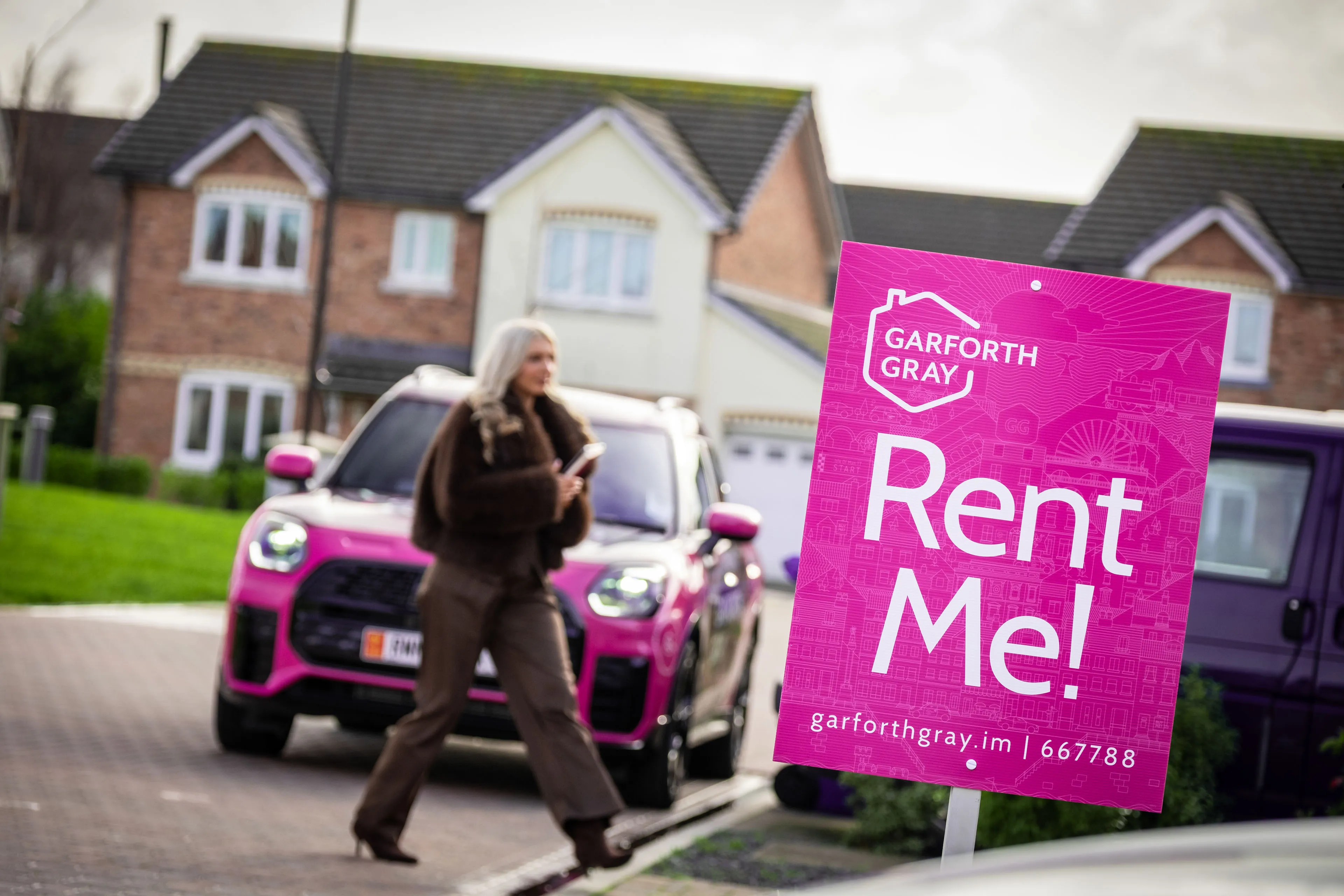 A "Rent Me!" sign from Garforth Gray is in focus. A woman walks with a phone in hand. Pink car and suburban houses in the background.