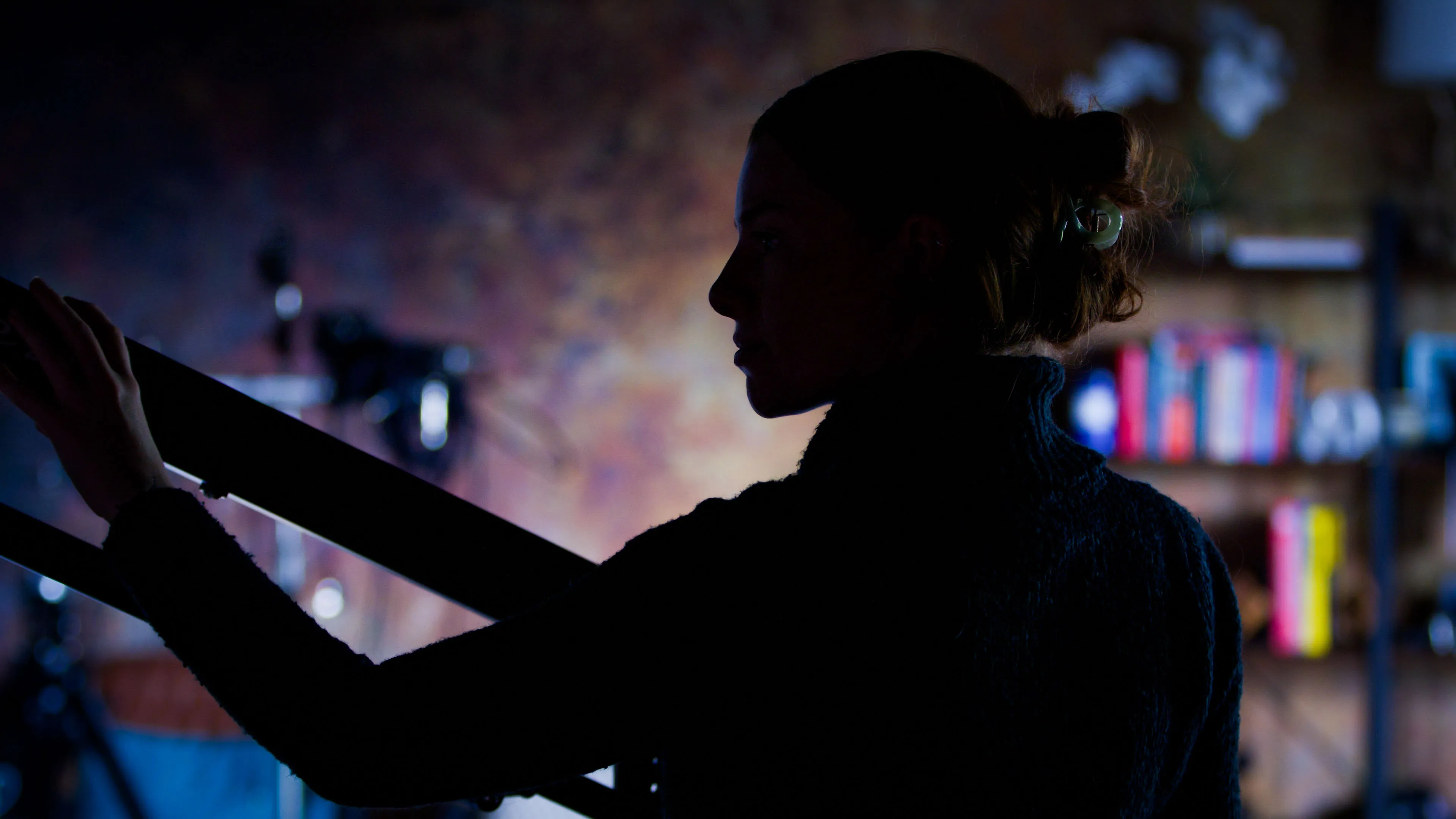 A silhouette of a woman in a dark room, holding a camera tripod. Dim lighting creates a moody atmosphere. Blurred bookshelf with colorful books in the background.