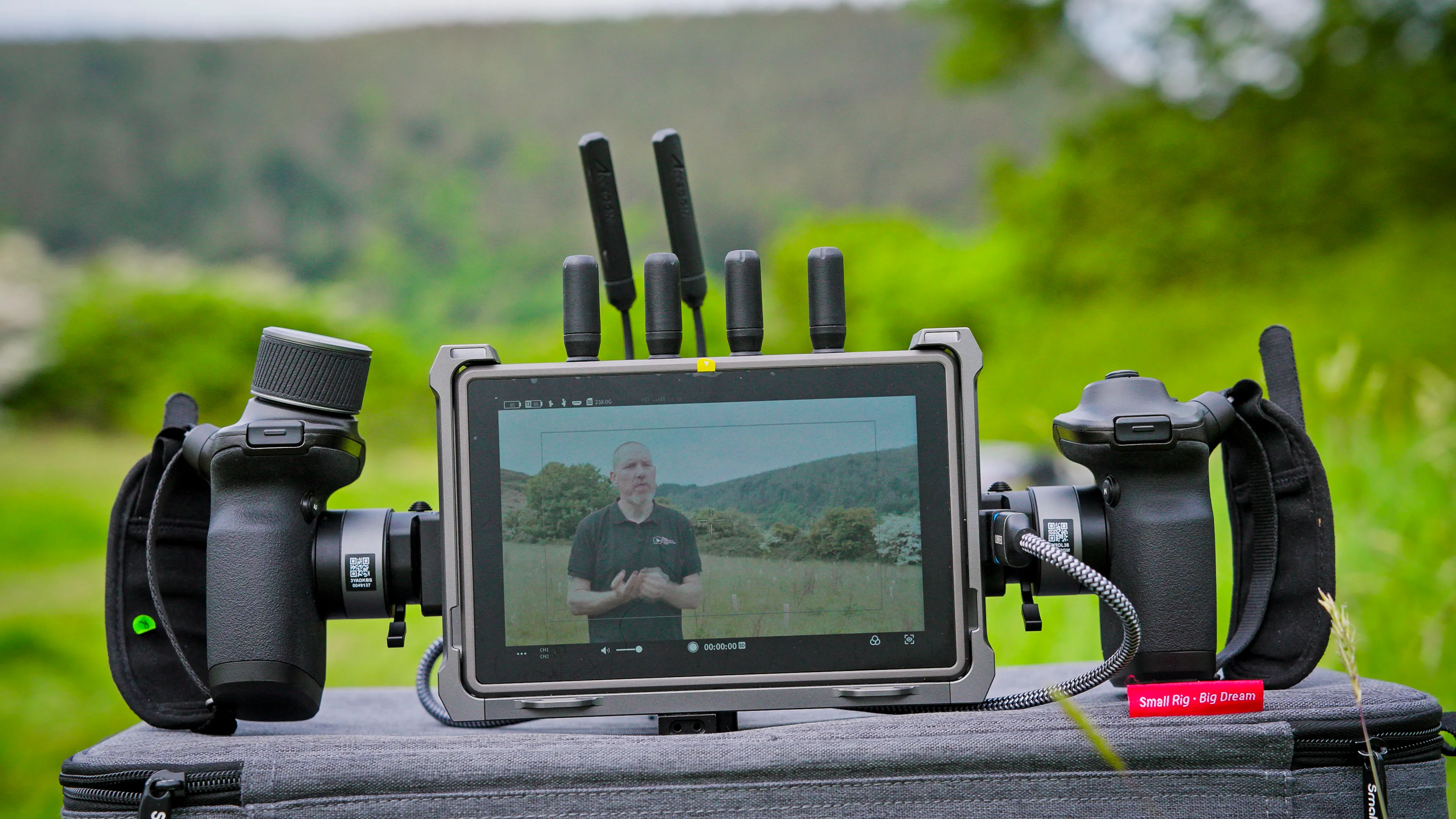 A rugged field monitor with dual handles displays a person speaking, set against a blurred, green outdoor backdrop.