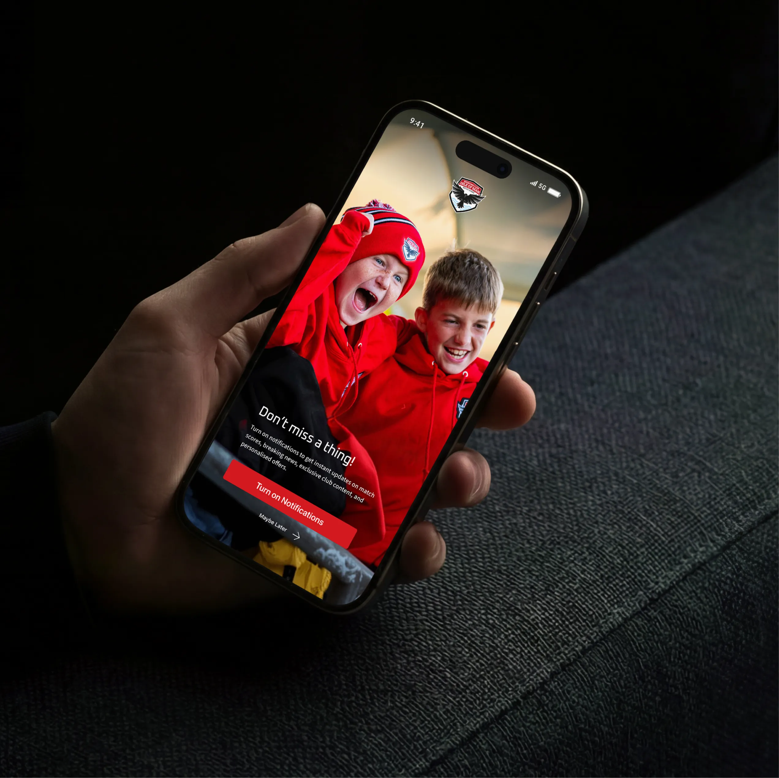 Hand holding a phone with the FC Isle of Man app asking supporters to turn on push notifications, above a photo of two young fans cheering