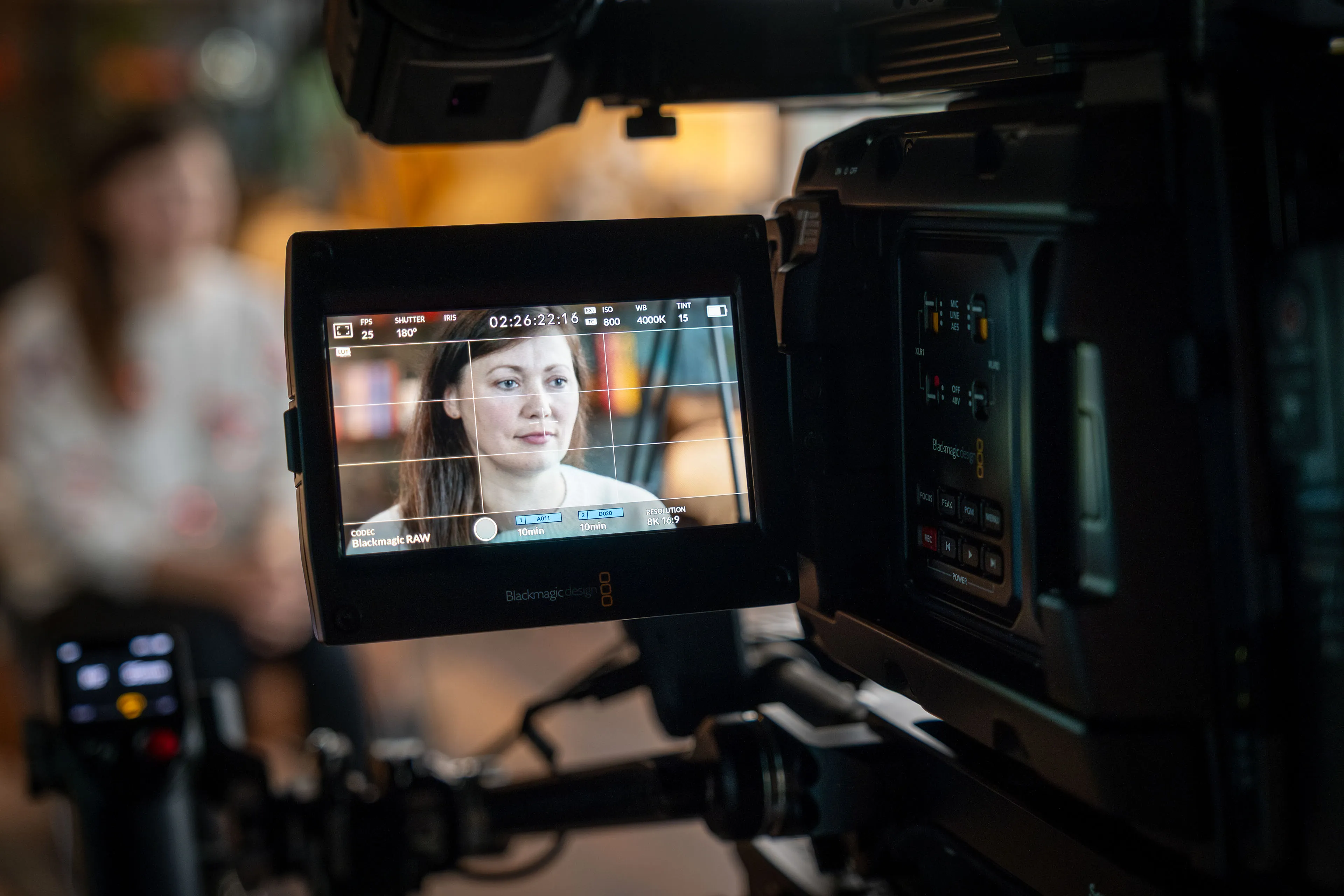 A professional camera captures a woman in focus on its screen, while she sits blurred in the background. The setting is warm and focused, conveying a serious tone.