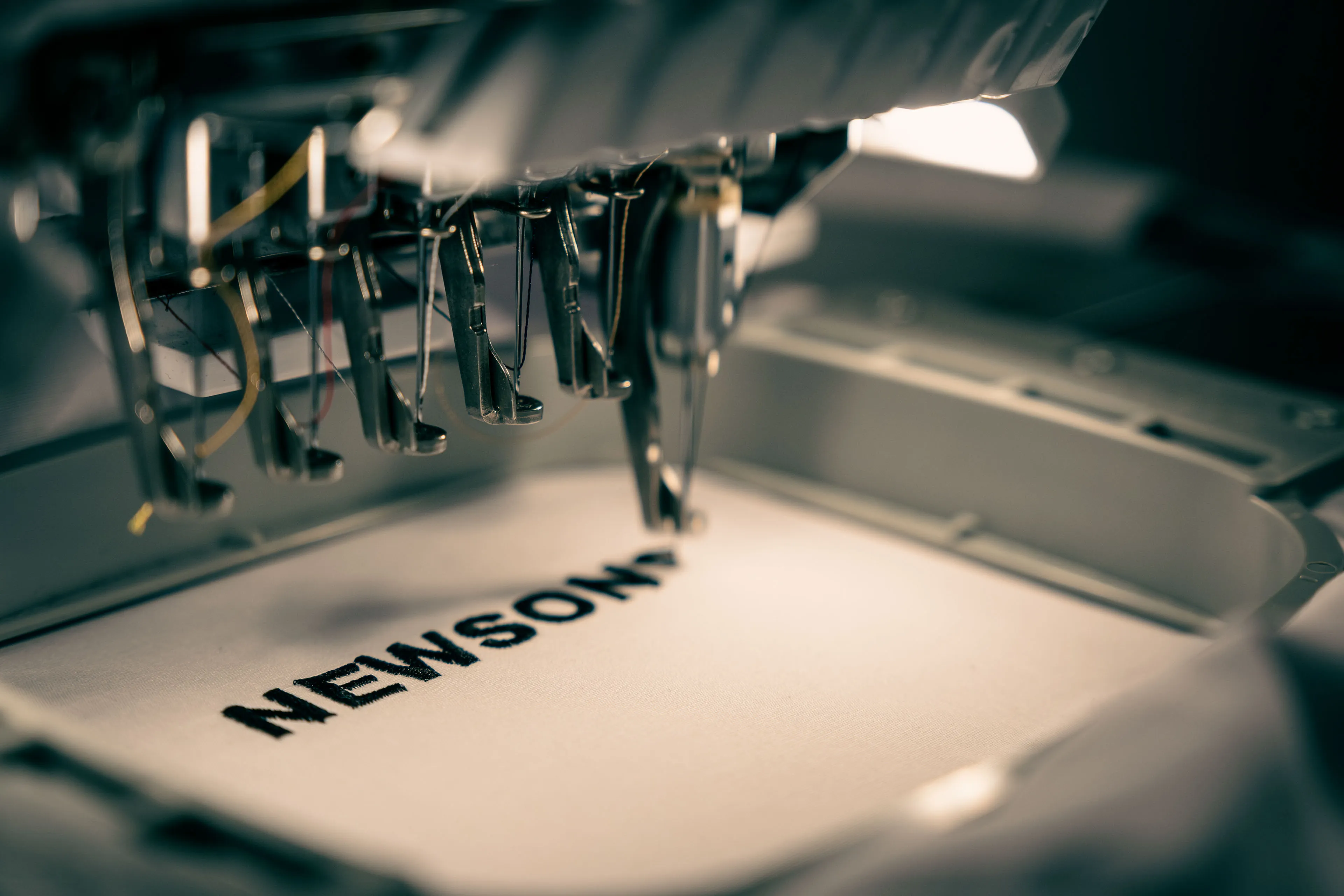 Close-up of an embroidery machine stitching the word "NEWSONS" on fabric. The scene is dimly lit, focusing on the machine's precision and detail.
