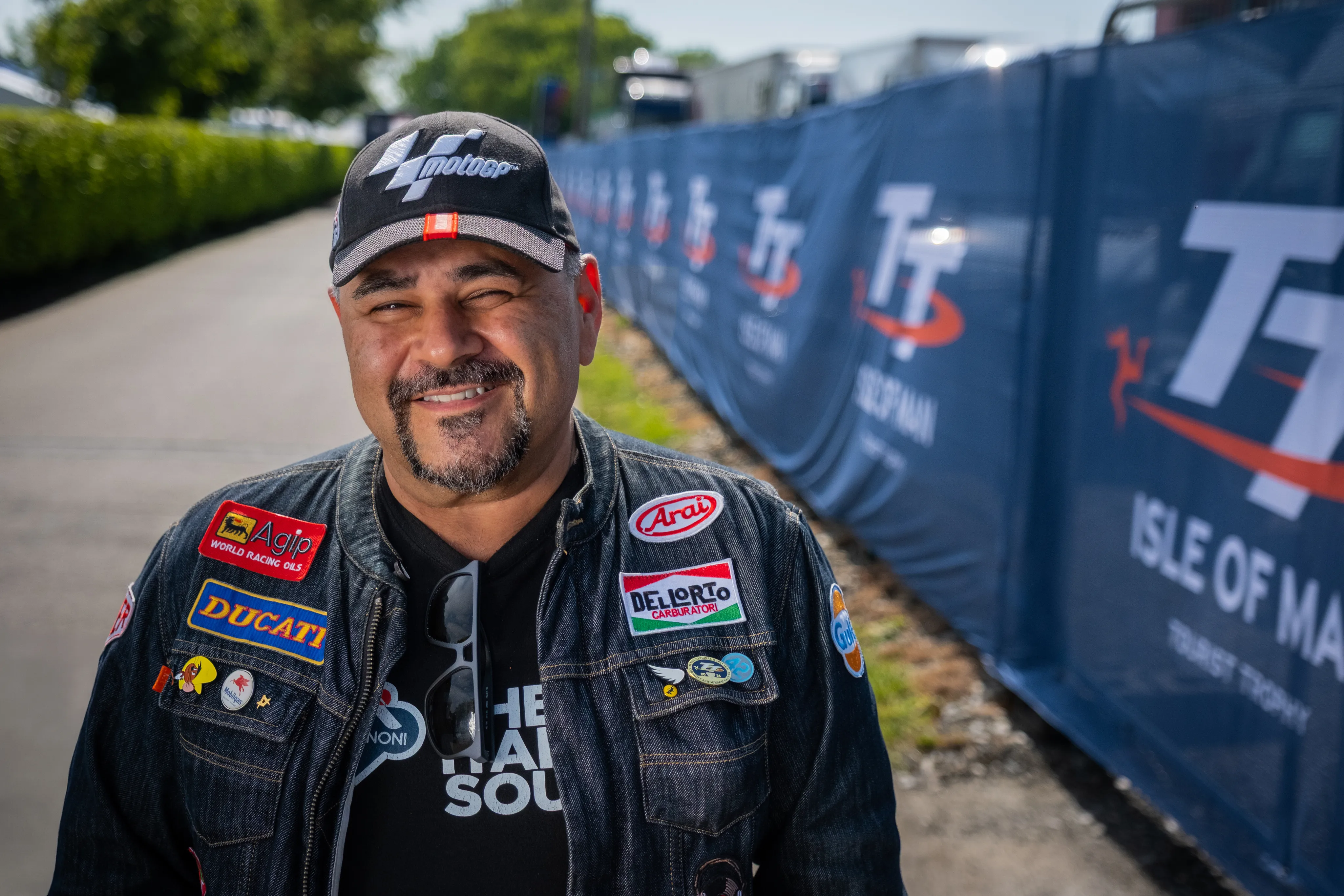 Ron Janjua from Brisbane, Australia, an Isle of Man TT Marshal, smiling outdoors for Behind the Tabard TT Marshals Series highlighting marshal stories, in a denim jacket full of patches