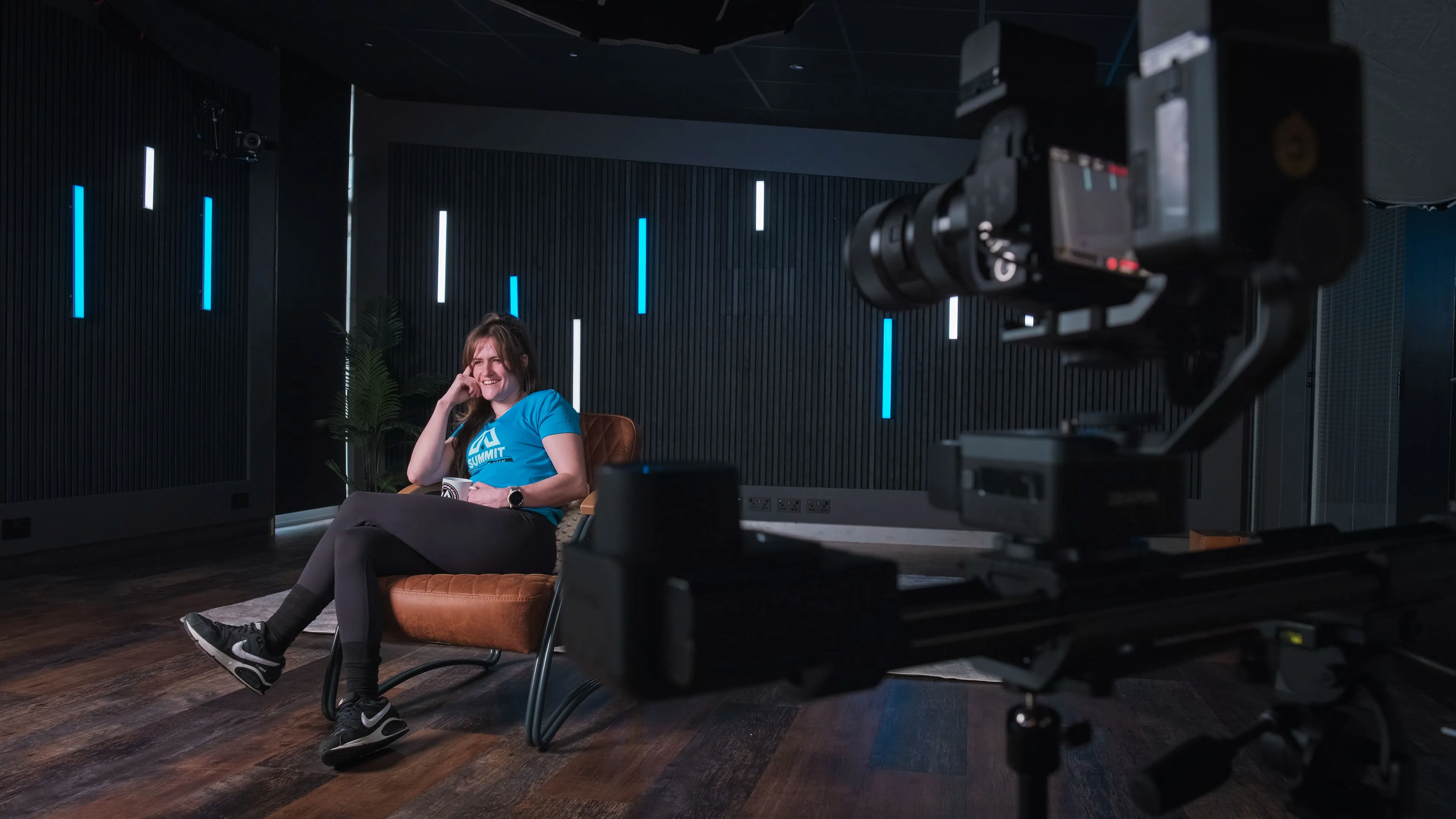 Woman in casual attire sits smiling on a chair in a dimly lit studio with vertical blue and white lights. Camera setup in the foreground. Relaxed atmosphere.