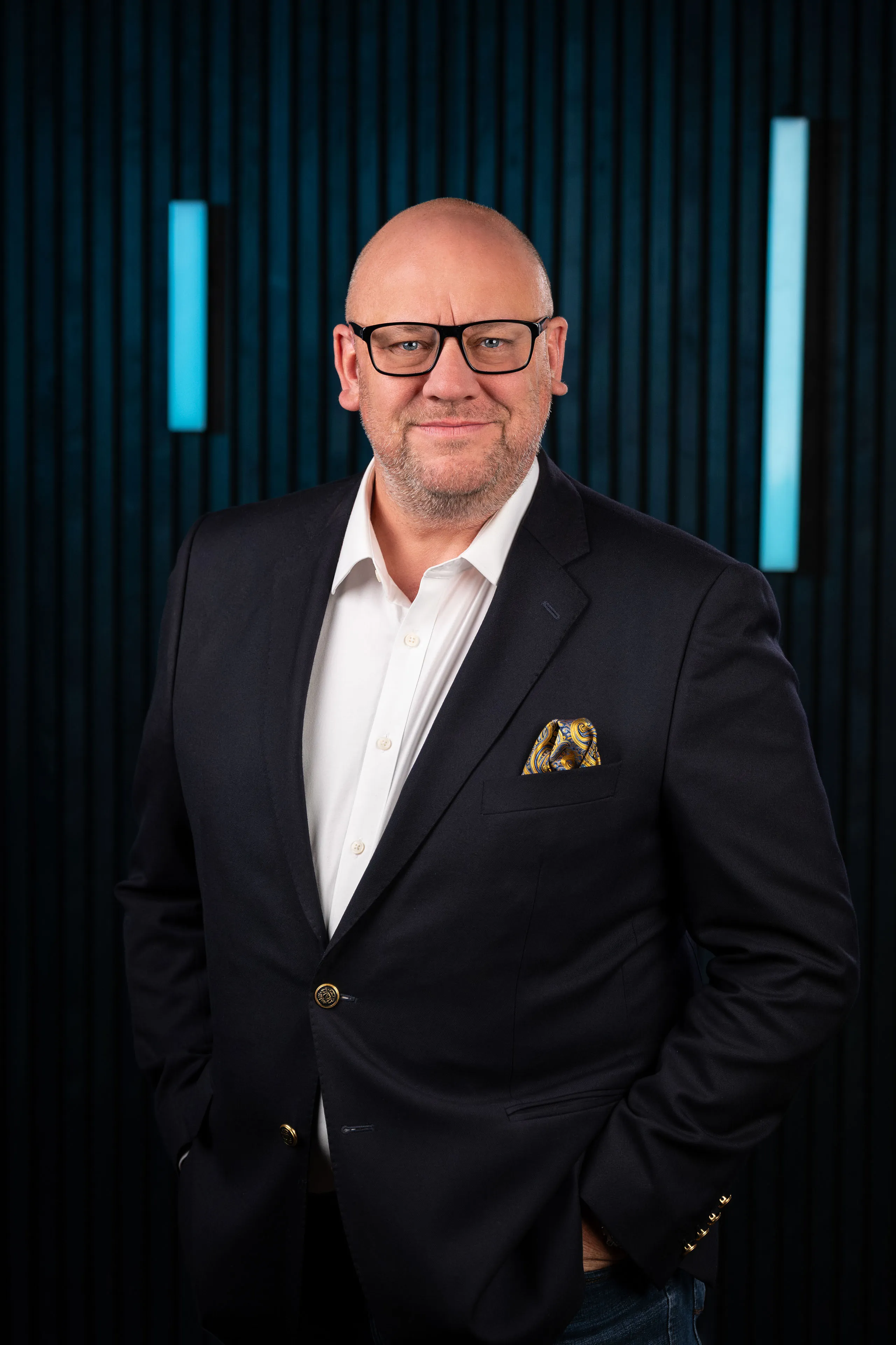 Professional headshot of Dave Bates wearing glasses and a dark blazer with a patterned pocket square, against a dark studio backdrop with blue accent lighting