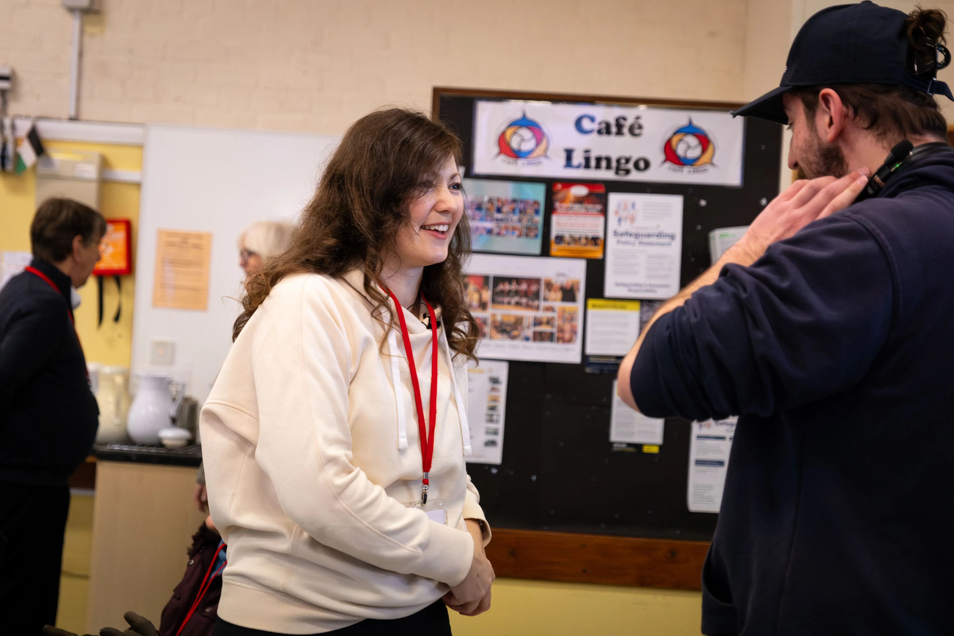 A woman in a white hoodie smiles while talking to a man in a dark cap in a community space with a "Café Lingo" sign and various posters on the wall, conveying a friendly, conversational atmosphere.