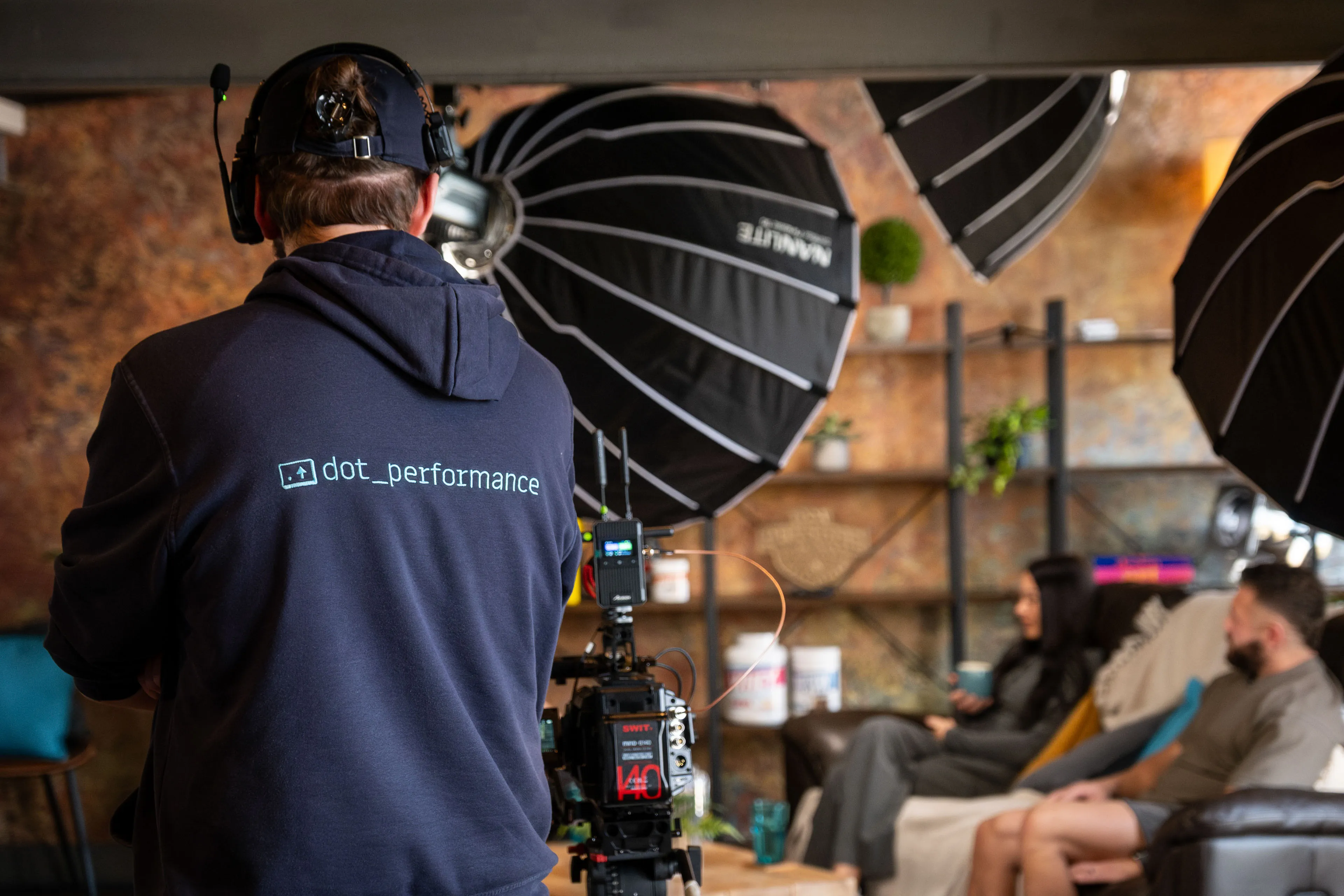 Will Oates wearing a DotPerformance branded hoodie operating a cinema camera during a podcast interview recording, with Danny and Christie Hands visible on a sofa in the background at DotPerformance studios on the Isle of Man