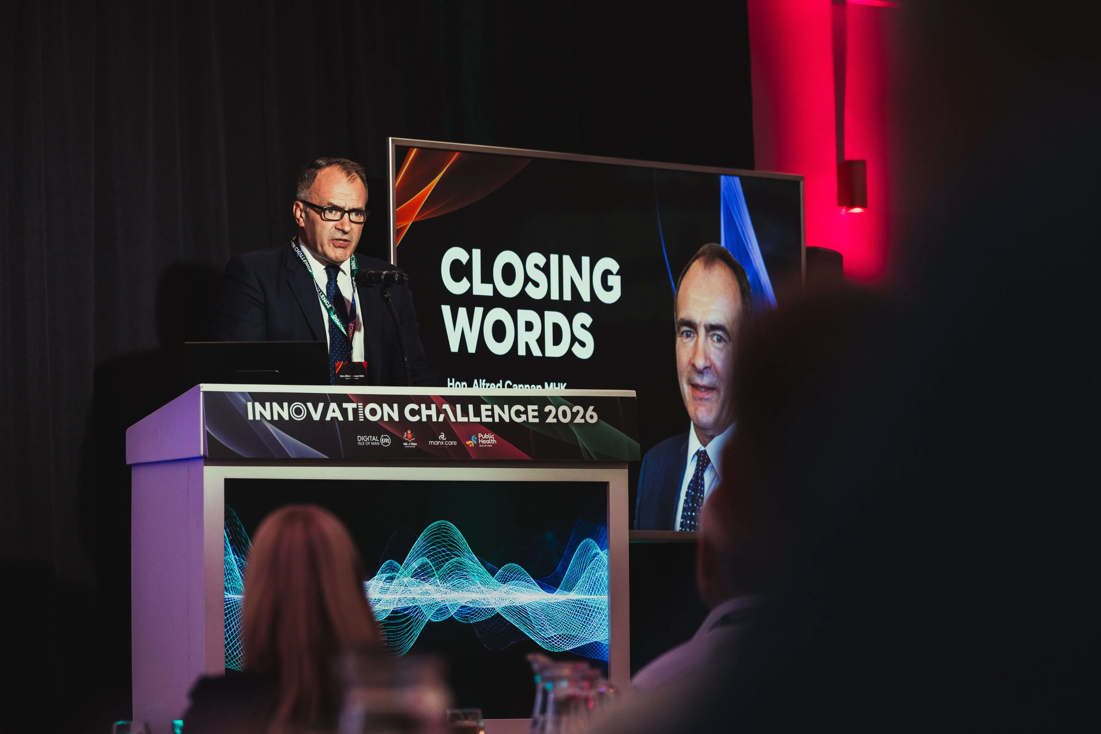 Hon. Alfred Cannan MHK speaking at the podium beside a "Closing Words" screen at the Innovation Challenge 2026