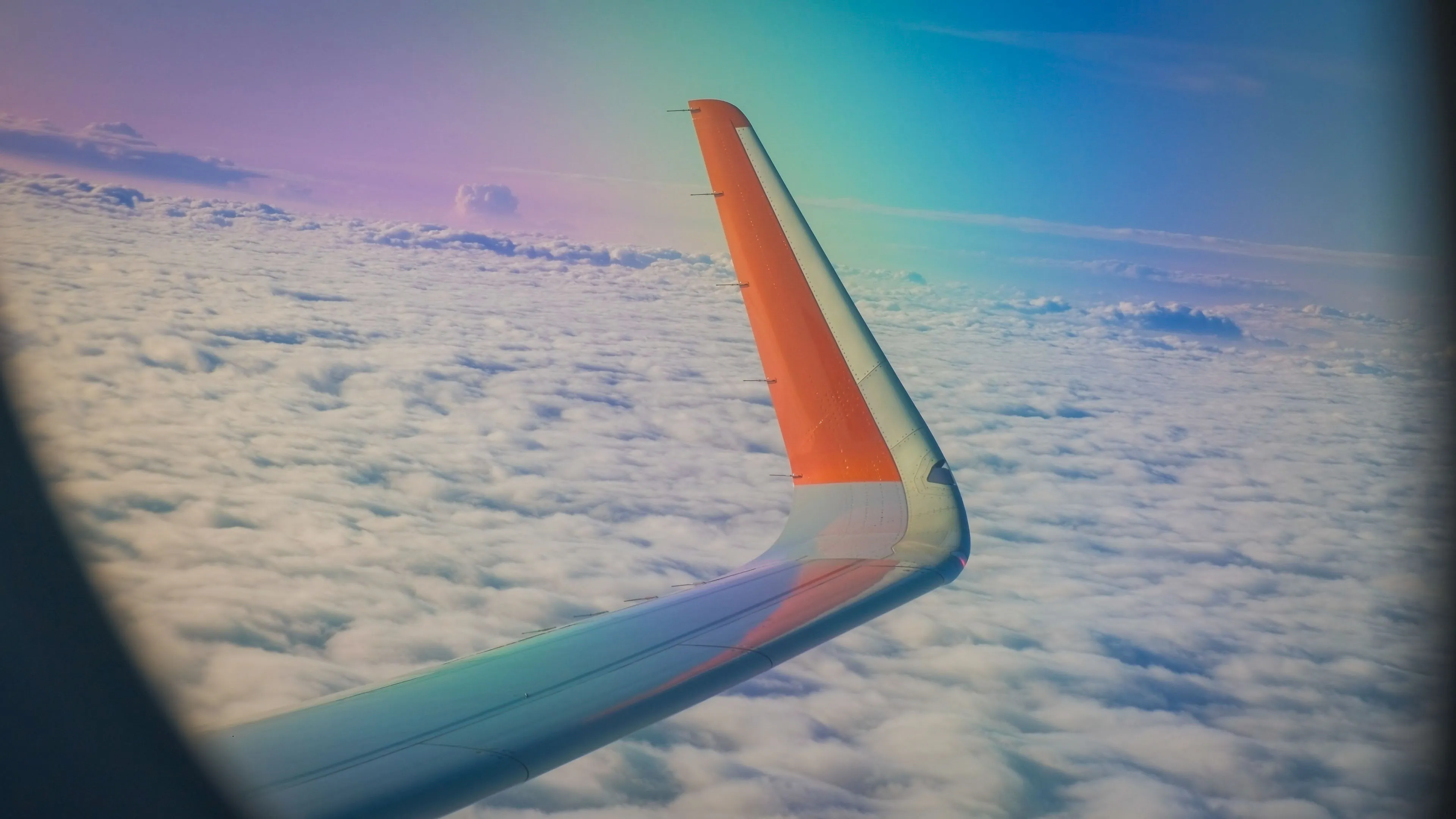 Airplane wing above fluffy white clouds at sunrise, with a gradient of pink, purple, and blue in the sky, creating a peaceful, dreamy atmosphere.