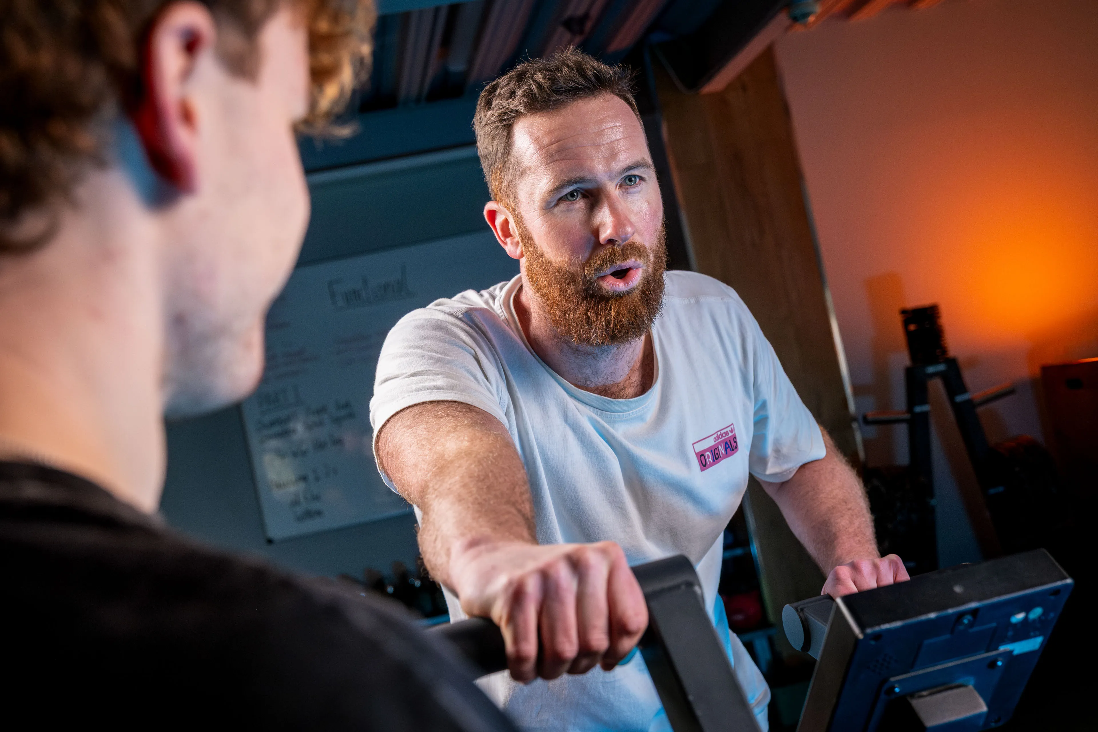 A person in a white shirt is focused on exercising at a gym, while a second individual is partially visible in the foreground.