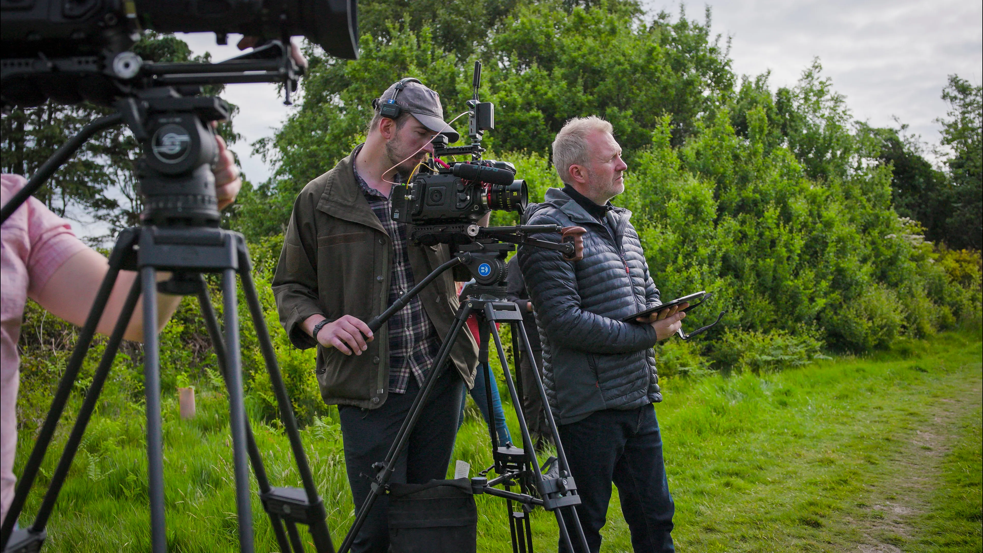 Two men stand outdoors with professional film cameras on tripods, surrounded by greenery. The scene conveys a focused and creative atmosphere.