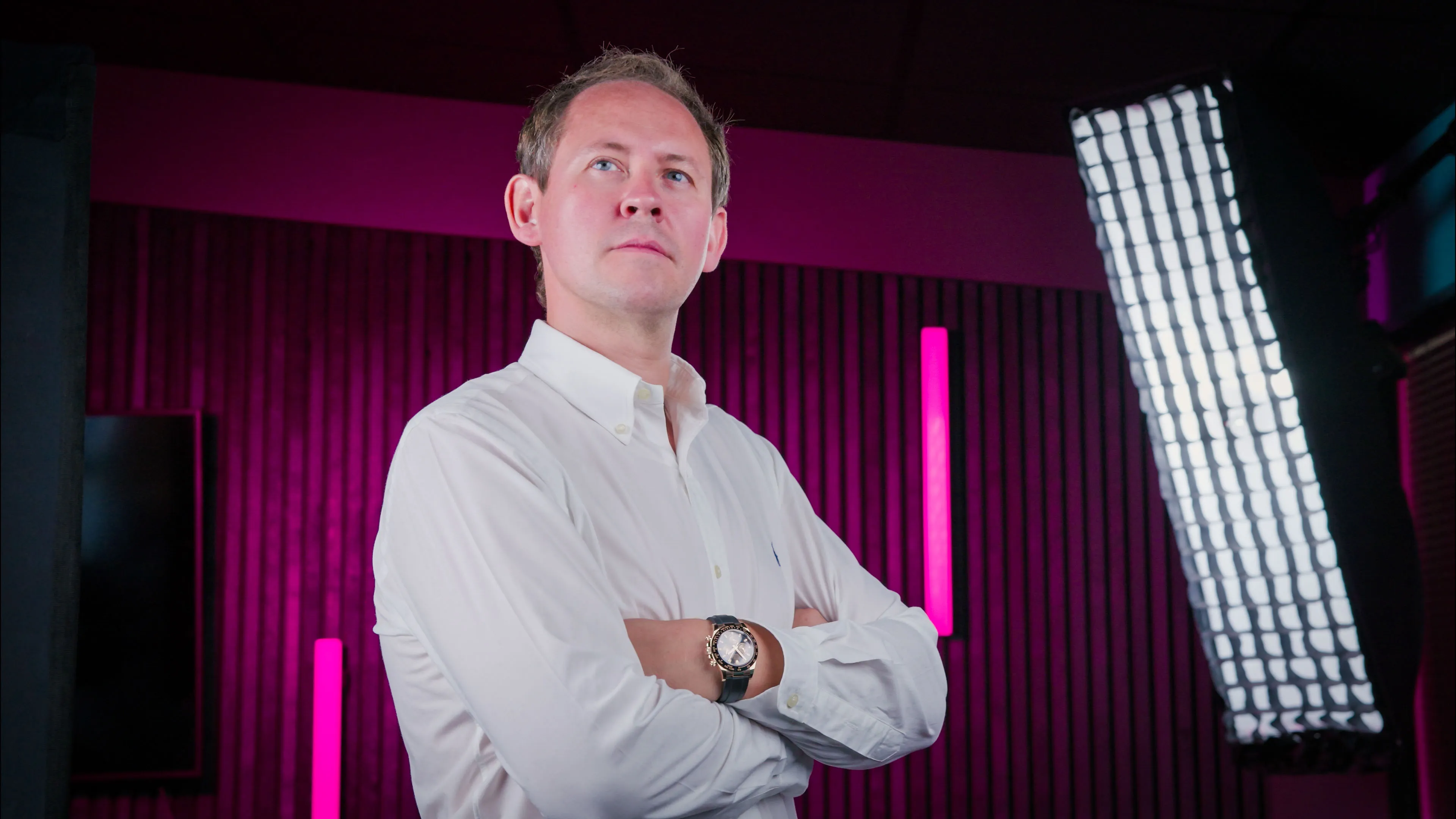 A person in a white shirt stands confidently in a studio with pink and purple lighting, looking upward. A large softbox light is visible on the right.