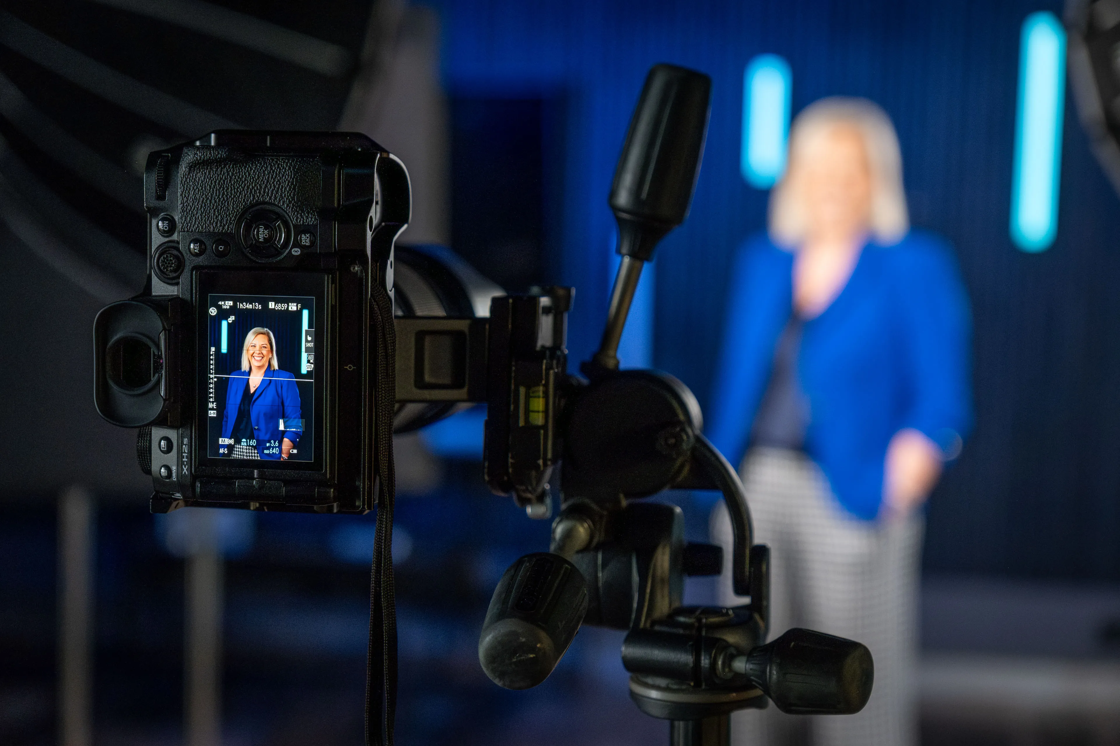 Camera screen showing a live preview of Lisa Karran's smiling headshot, with Lisa blurred in the background against blue studio lighting