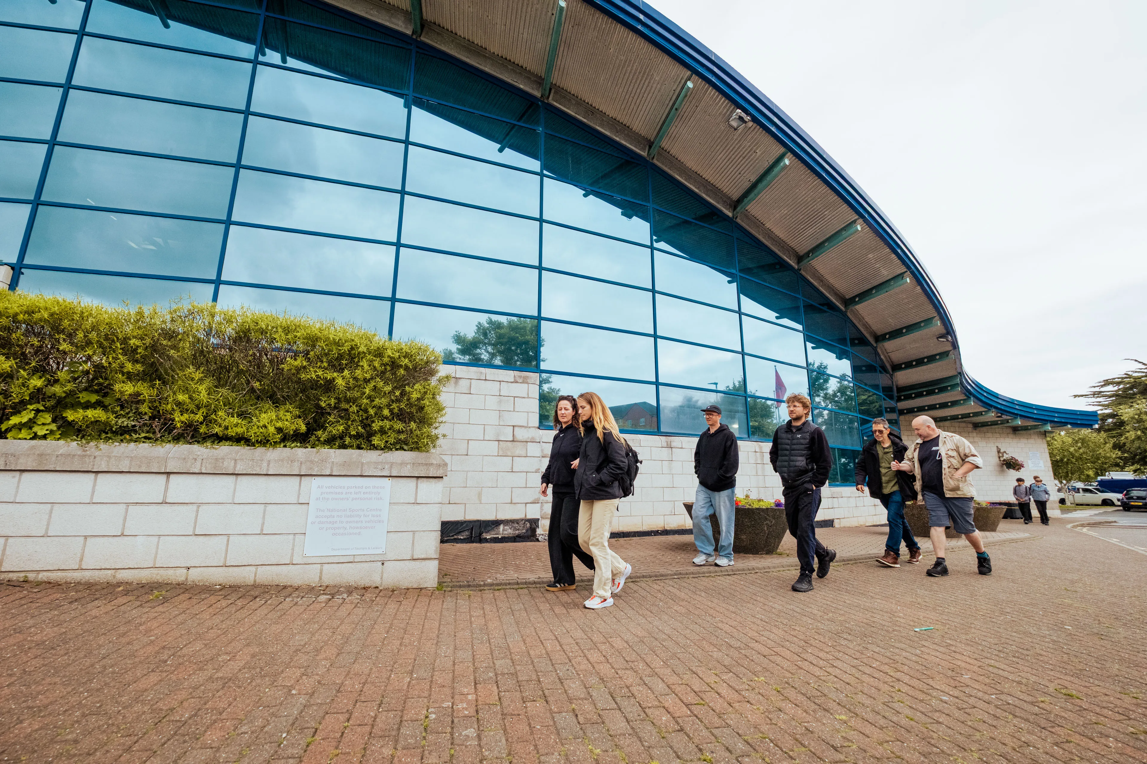 A group of people walk in front of a modern building with large blue-tinted windows and a curved roof. The mood is casual and relaxed.