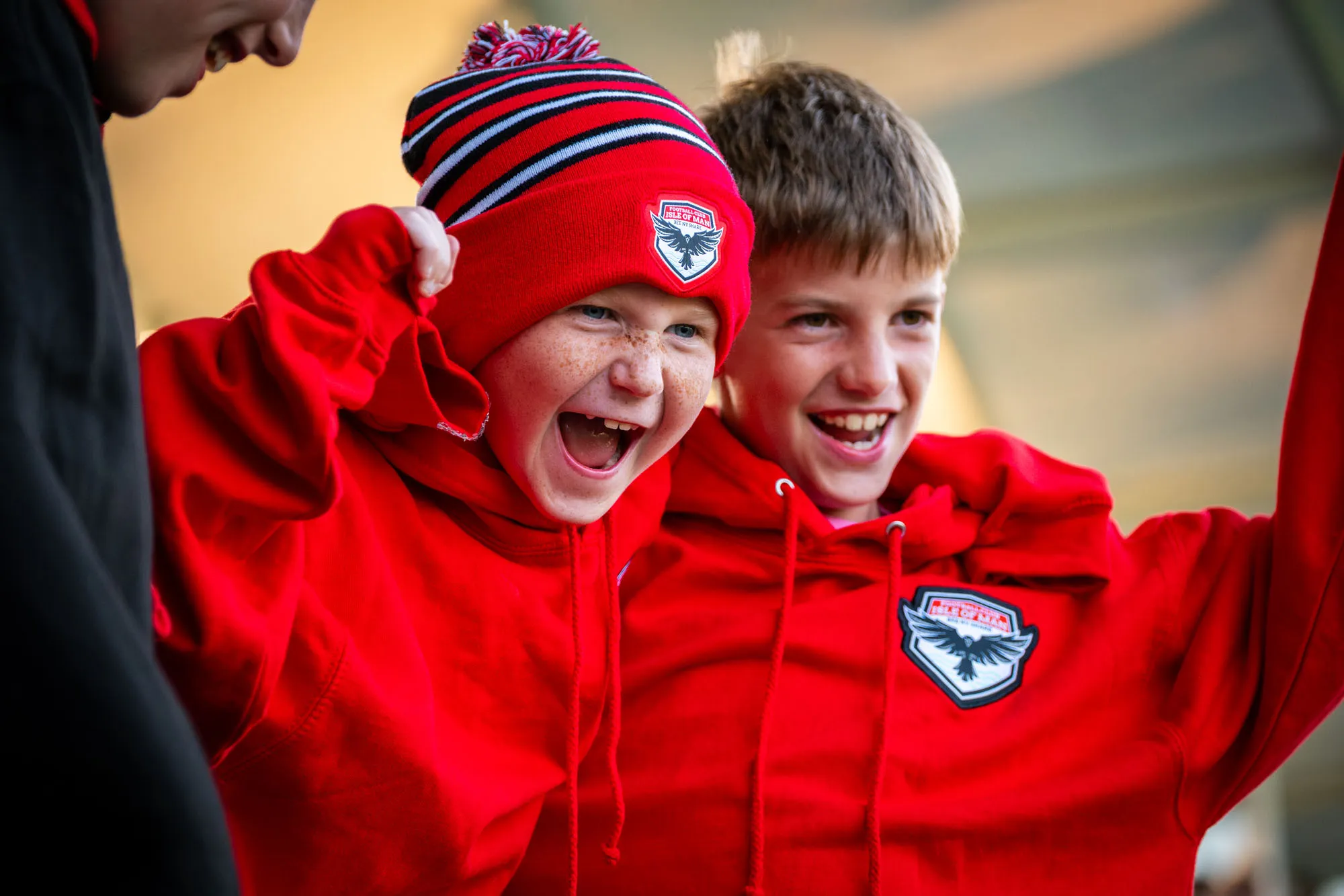 Two young supporters laughing together while wearing Ravens beanie and hoodie merch at an FCIOM match