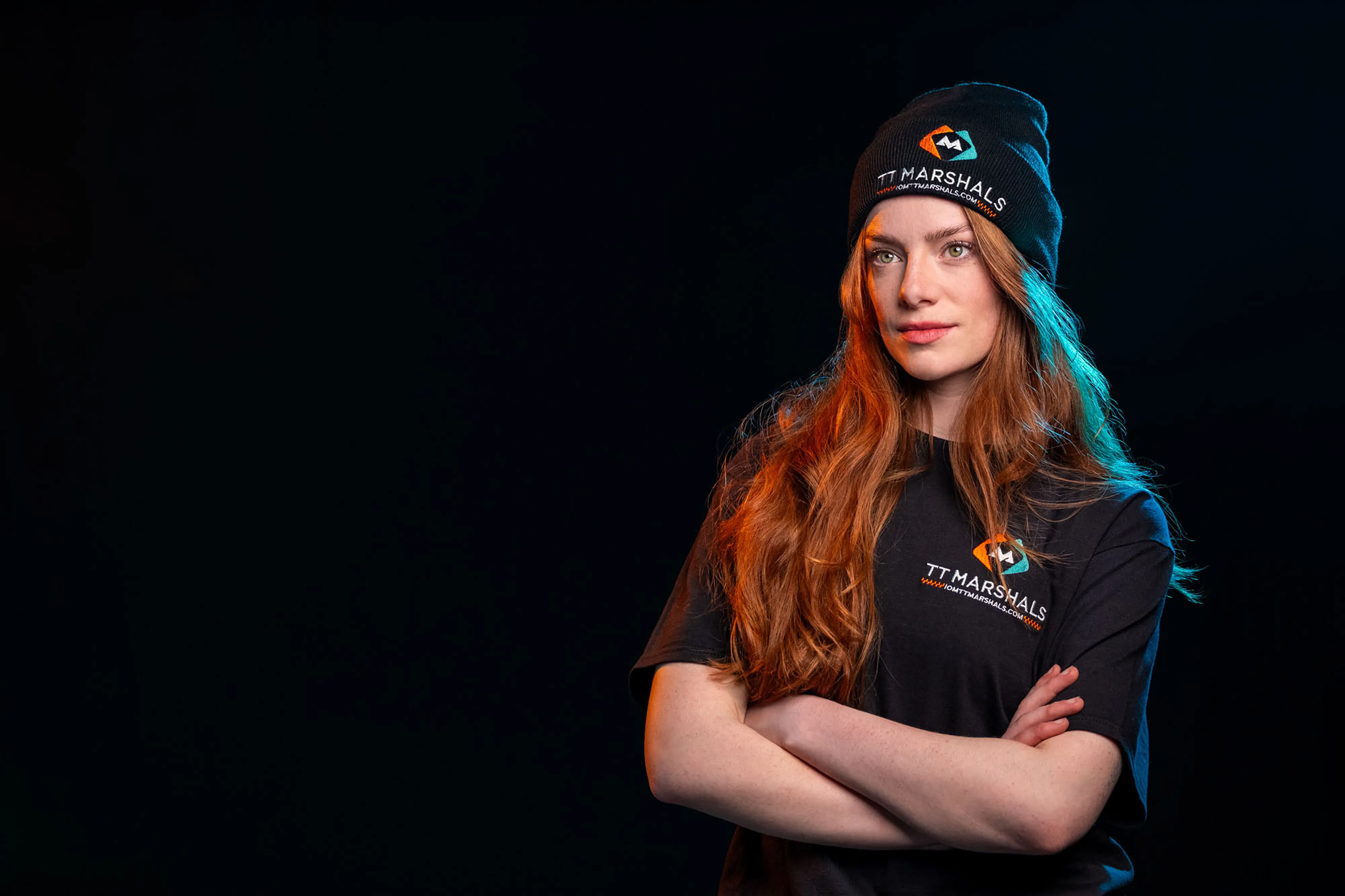 Ella Garvey in Marshals Merchandise, exclusively for marshals for TT 2026. Ella poses with crossed arms and looks away from the camera, set against a black background, wearing a marshals beanie and t-shirt.