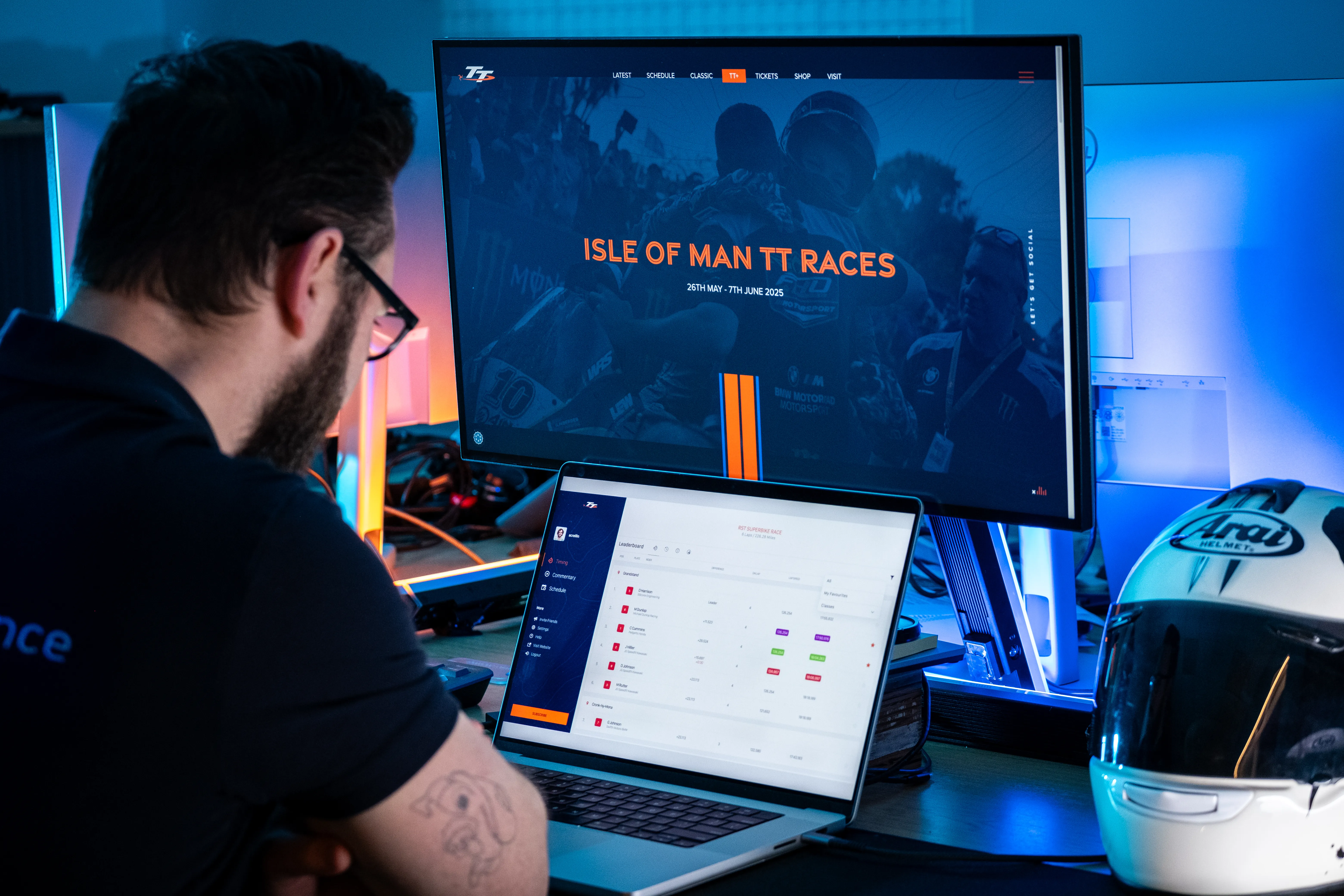 A DotPerformance developer working at a multi-monitor setup displaying the Isle of Man TT Races website and the DotPerformance live timing platform leaderboard, with an Arai helmet visible on the desk