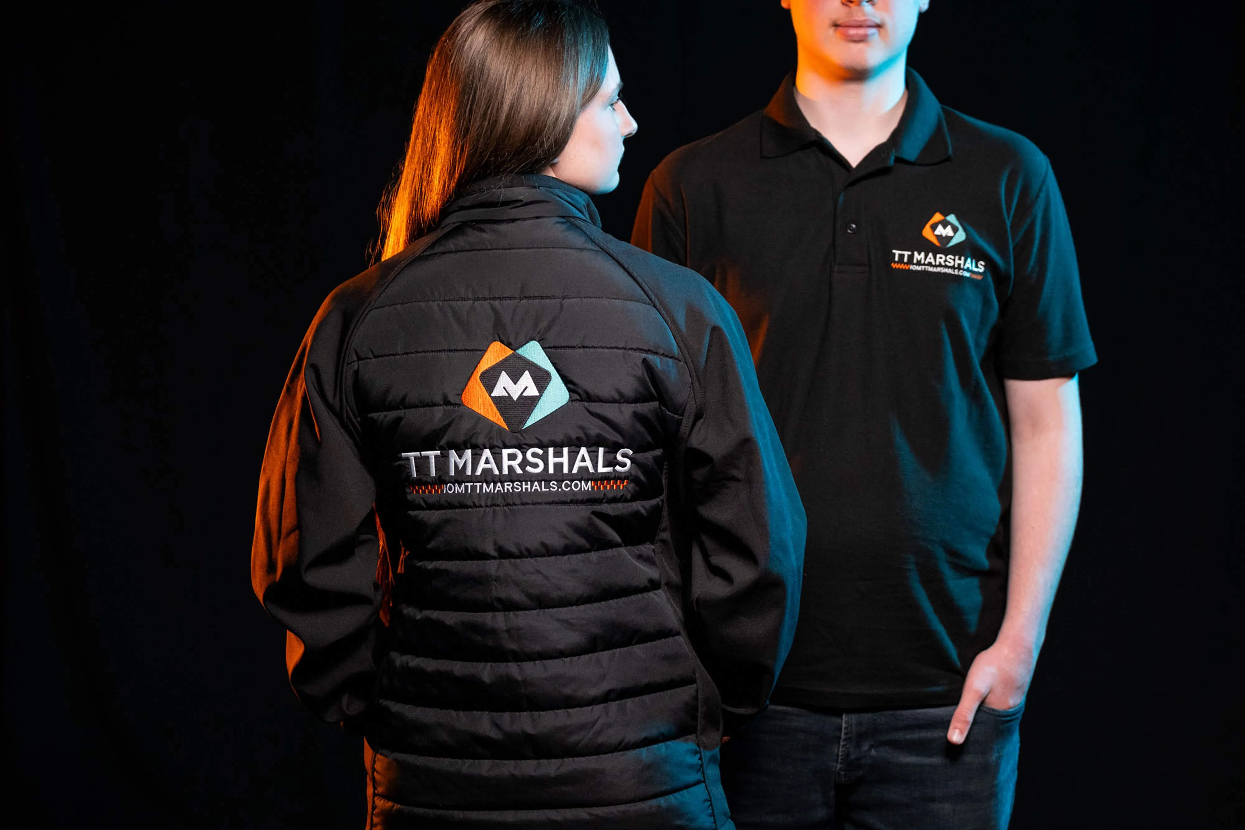 Caitlin Hampson and Deane Corlett Aspell wearing the official TT Marshals 2026 black polo shirt and softshell padded jacket, featuring the TT Marshals logo. Caitlin with her back to the camera. Exclusive marshal merchandise for the Isle of Man TT 2026.