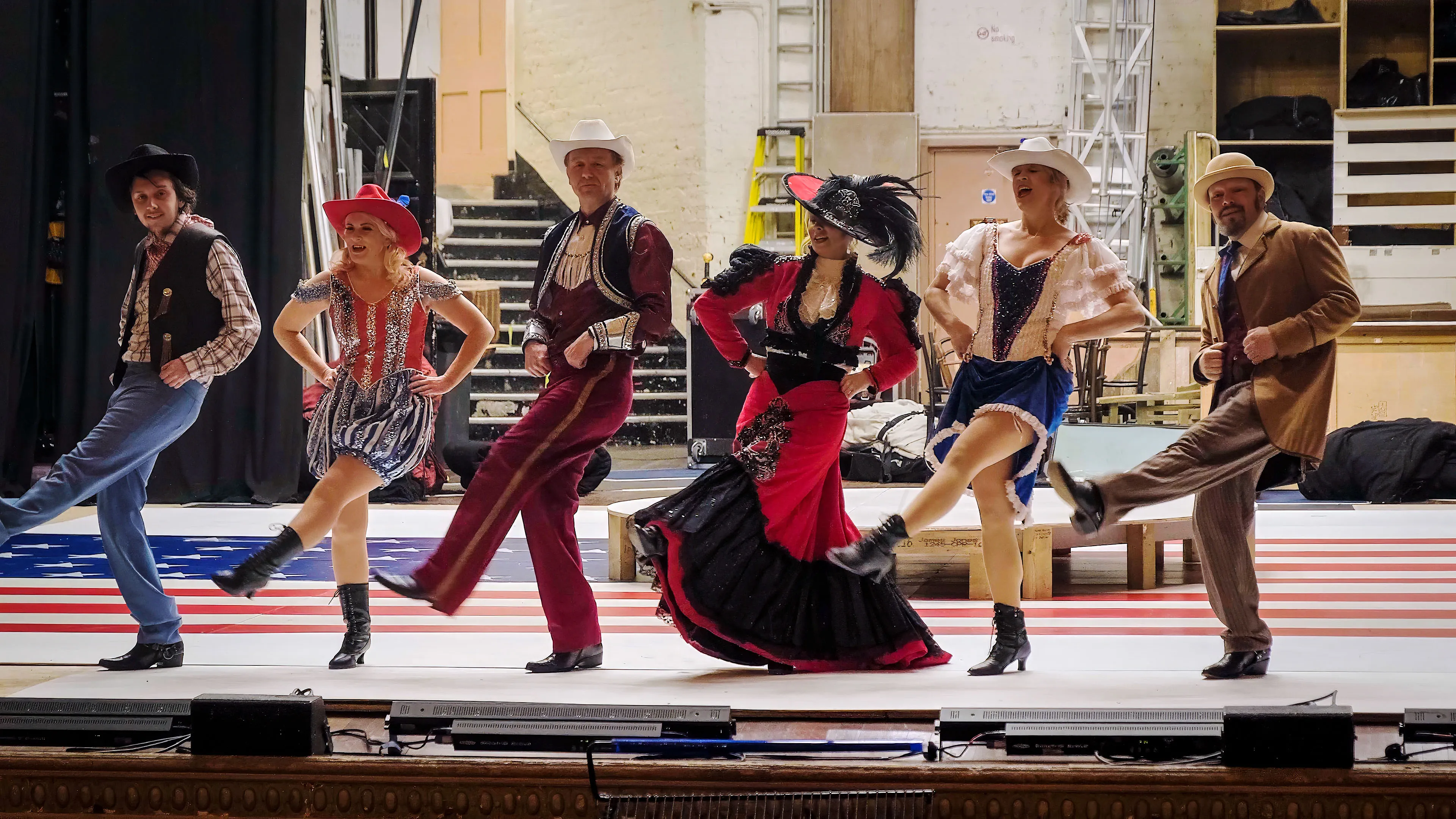 Six performers in colorful Western costumes dance in unison on stage, with an American flag pattern in the background. They appear joyful and lively.