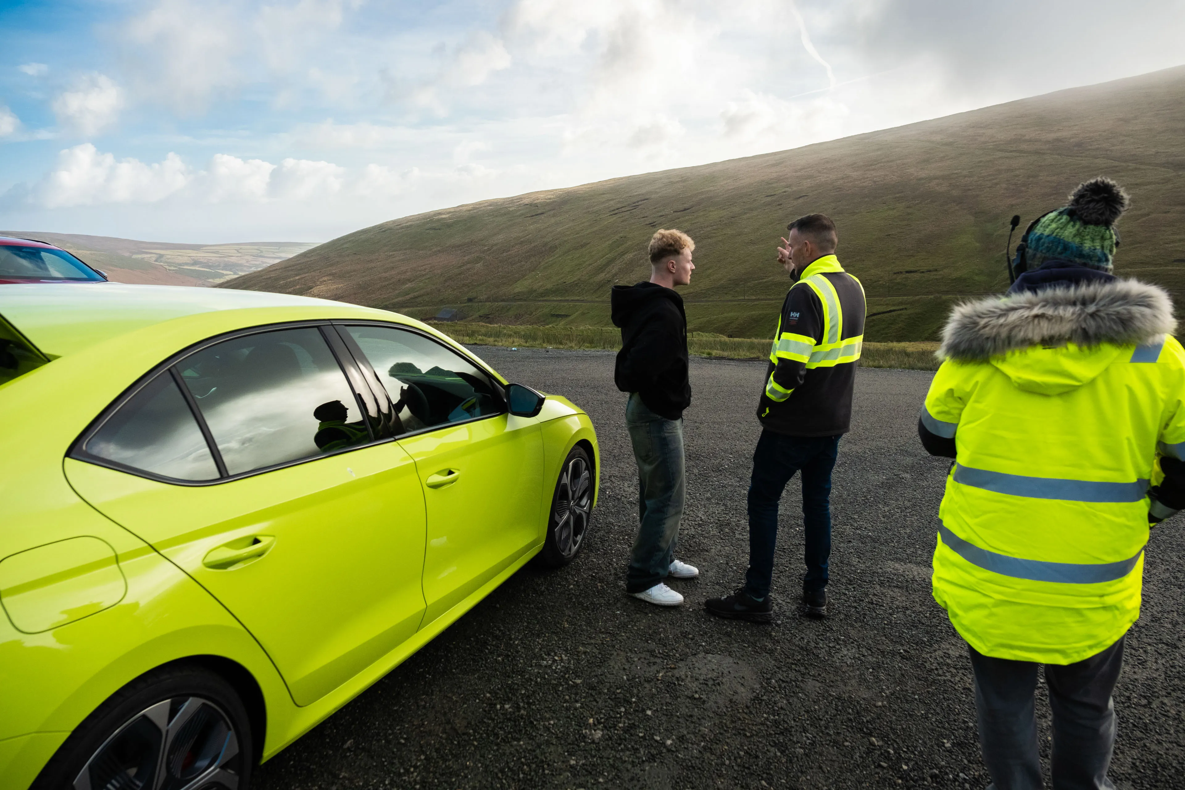 Two men stand beside a bright lime green Skoda car, engaged in conversation. Another person in a high-vis jacket looks on. Rolling hills are visible in the background under a cloudy sky.