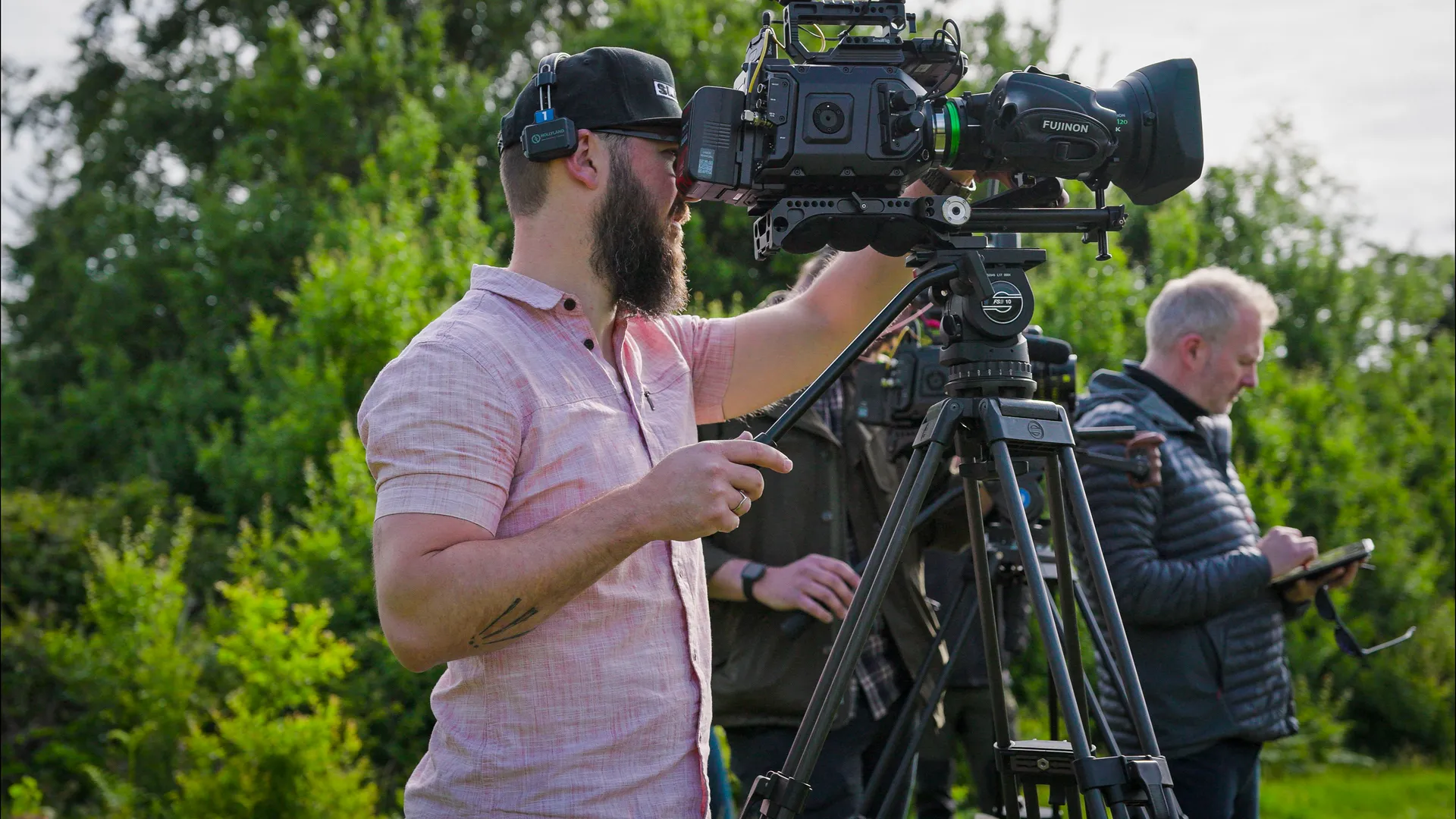 A bearded man in a red shirt operates a professional camera on a tripod, focusing intently. In the background, another person takes notes on a tablet. The scene is set outdoors with lush greenery, conveying a focused and diligent atmosphere.