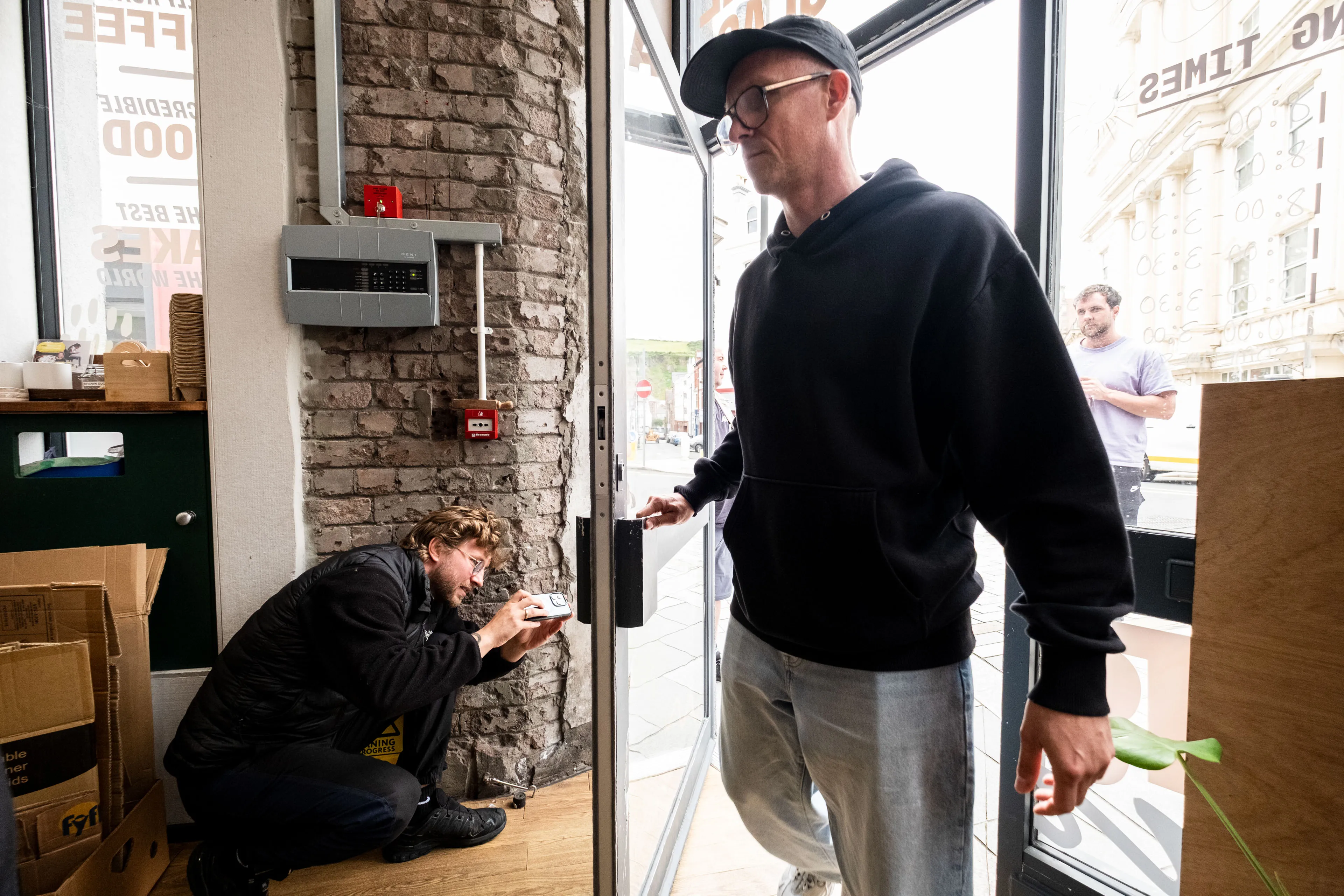 A man in a black hoodie enters a café, while another man crouches nearby with a camera. The scene is casual and candid, with a relaxed and curious tone.