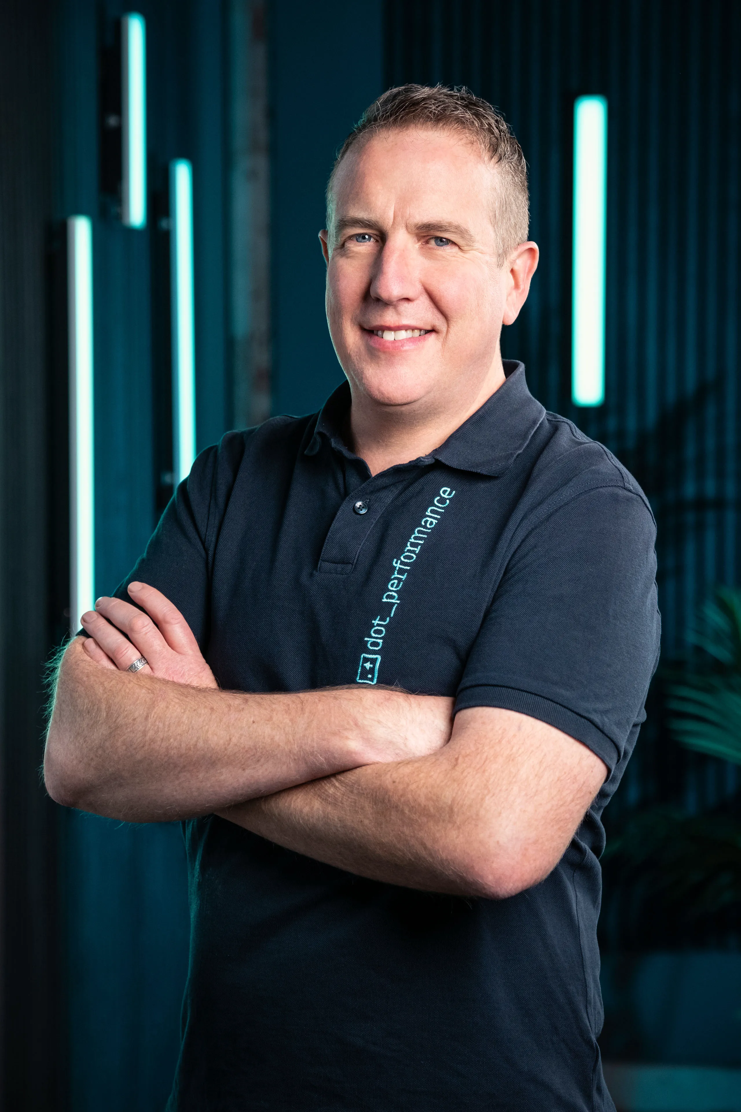 Simon King, Head of Systems at DotPerformance studios on the Isle of Man, smiling with arms crossed wearing a DotPerformance branded polo shirt, against a dark background with teal neon lighting.