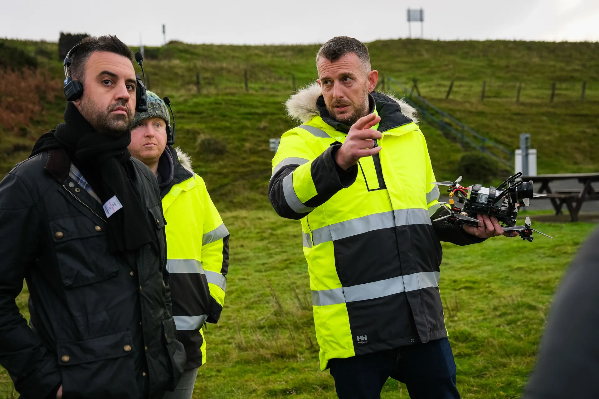 Three men in bright safety jackets stand on grass, with one pointing and holding a drone. The setting seems calm and focused, outdoors on a hill.