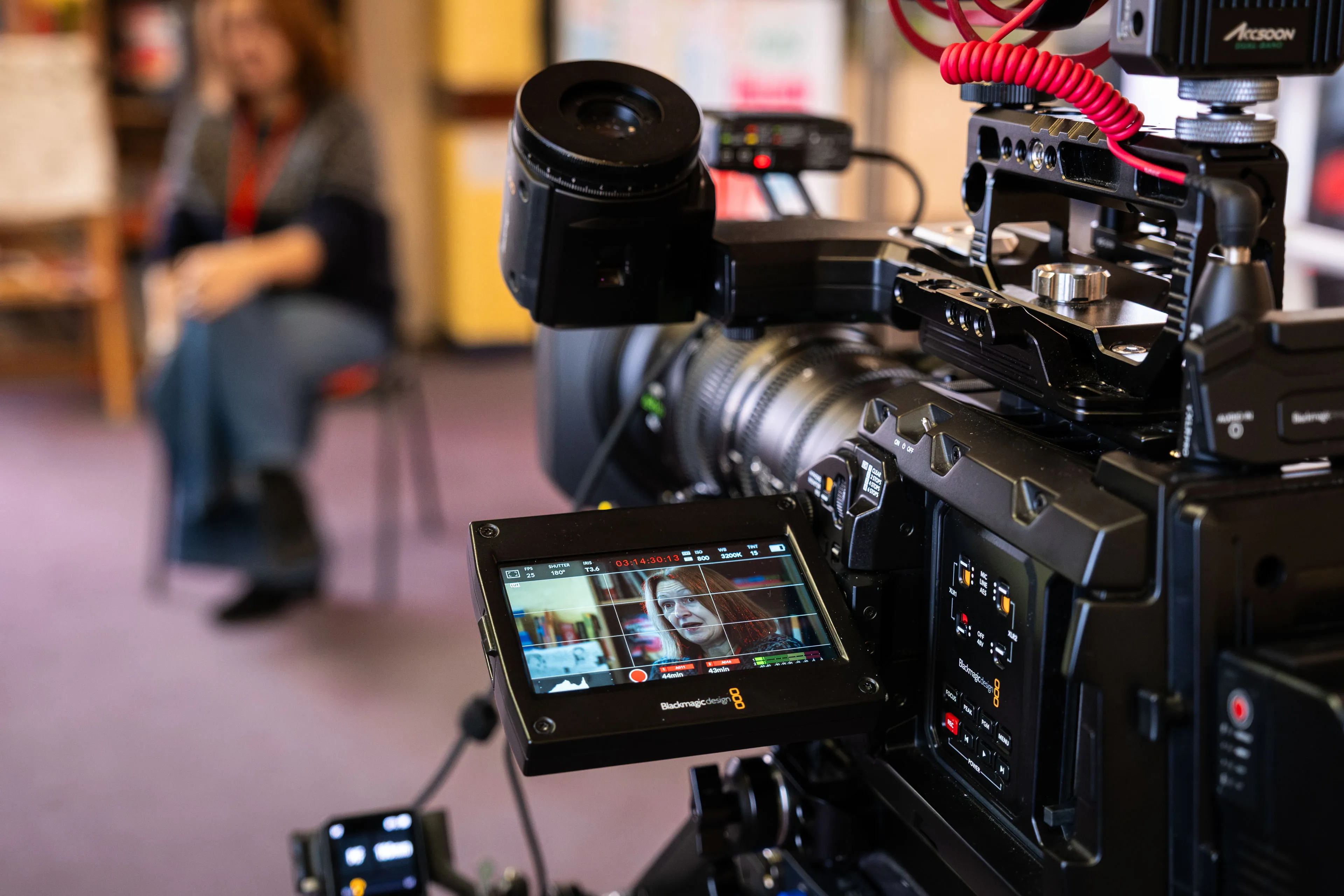 A professional video camera focuses on a blurred person sitting indoors. The camera's monitor shows a clear image of the seated individual.