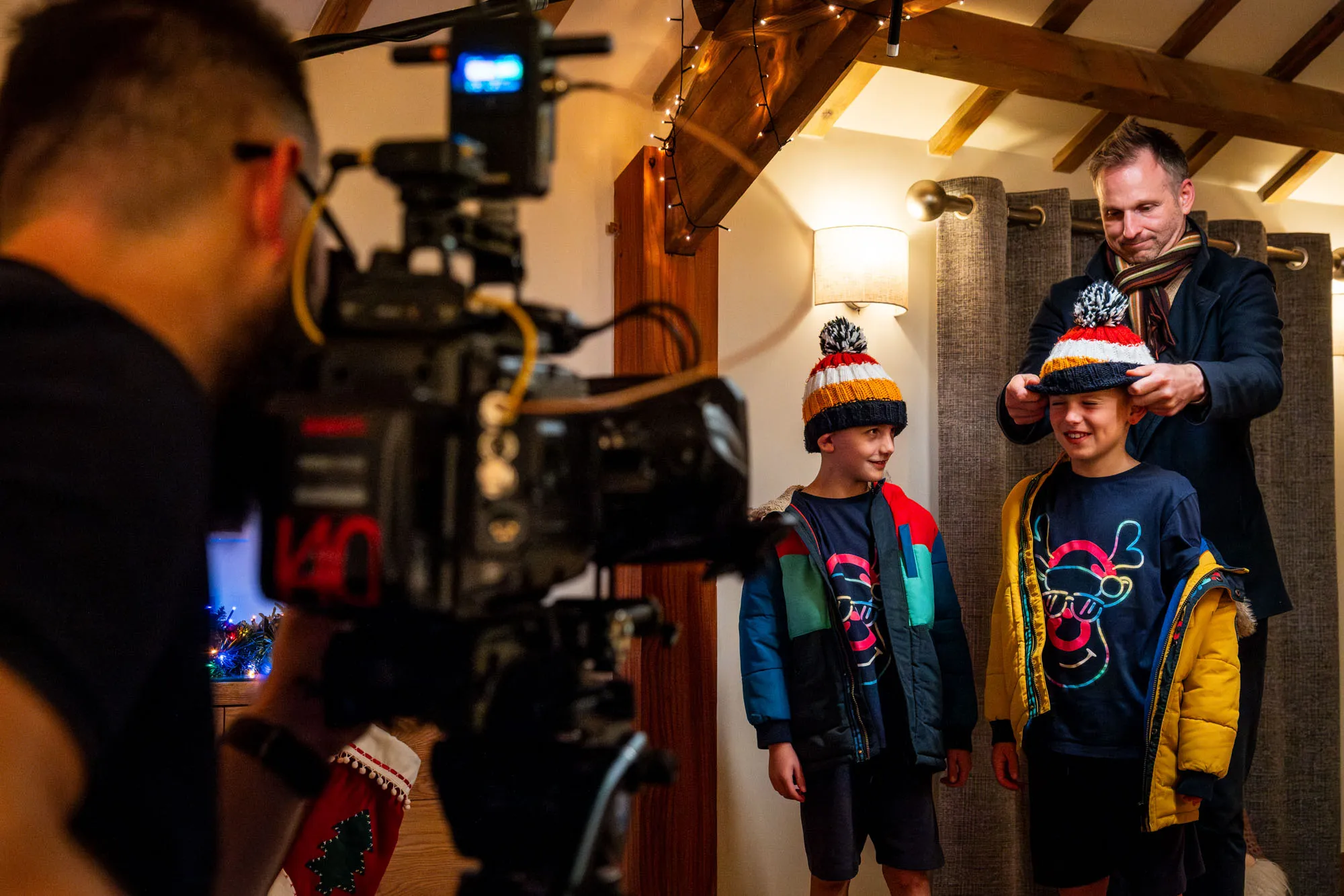 A man adjusts a colourful winter hat on a smiling boy, next to another boy in similar attire. A cameraman films them in a warmly lit room.