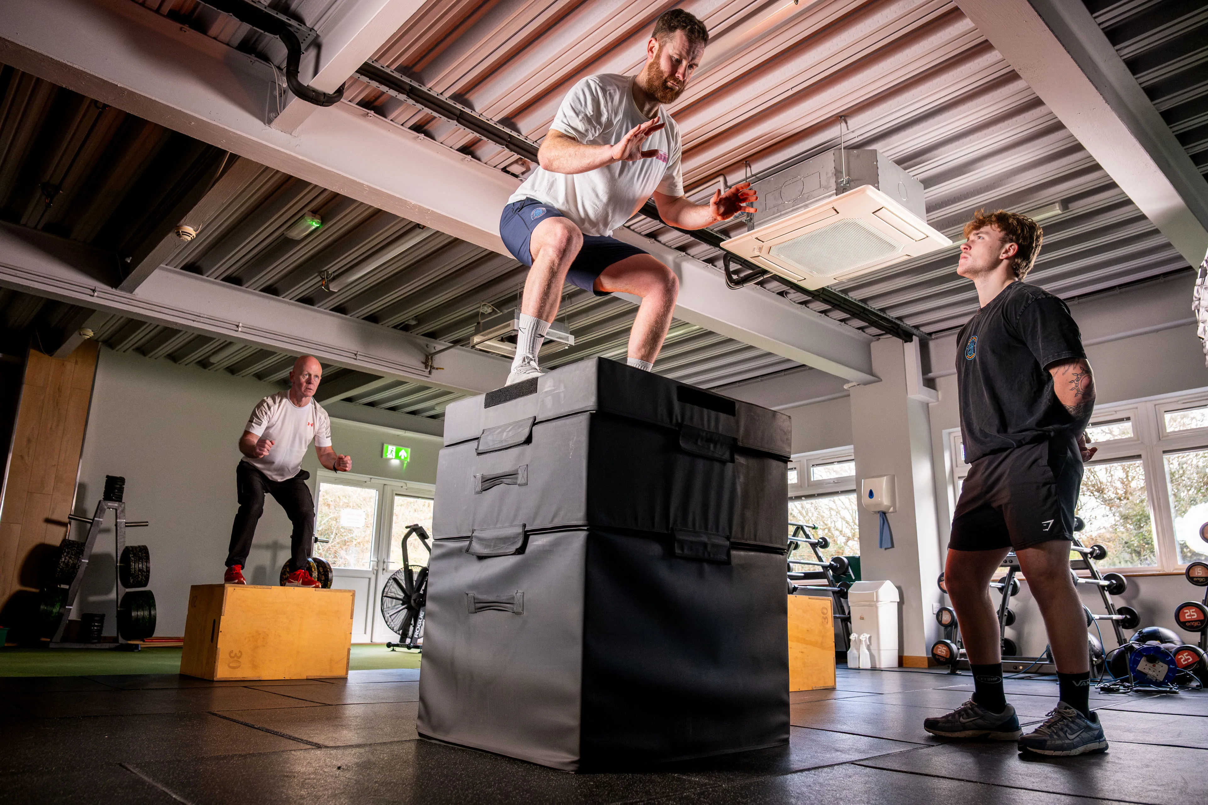 Two men exercise in a gym; one performs a box jump onto a high platform, while another supervises. A third person jumps onto a smaller box. The atmosphere is focused and energetic.