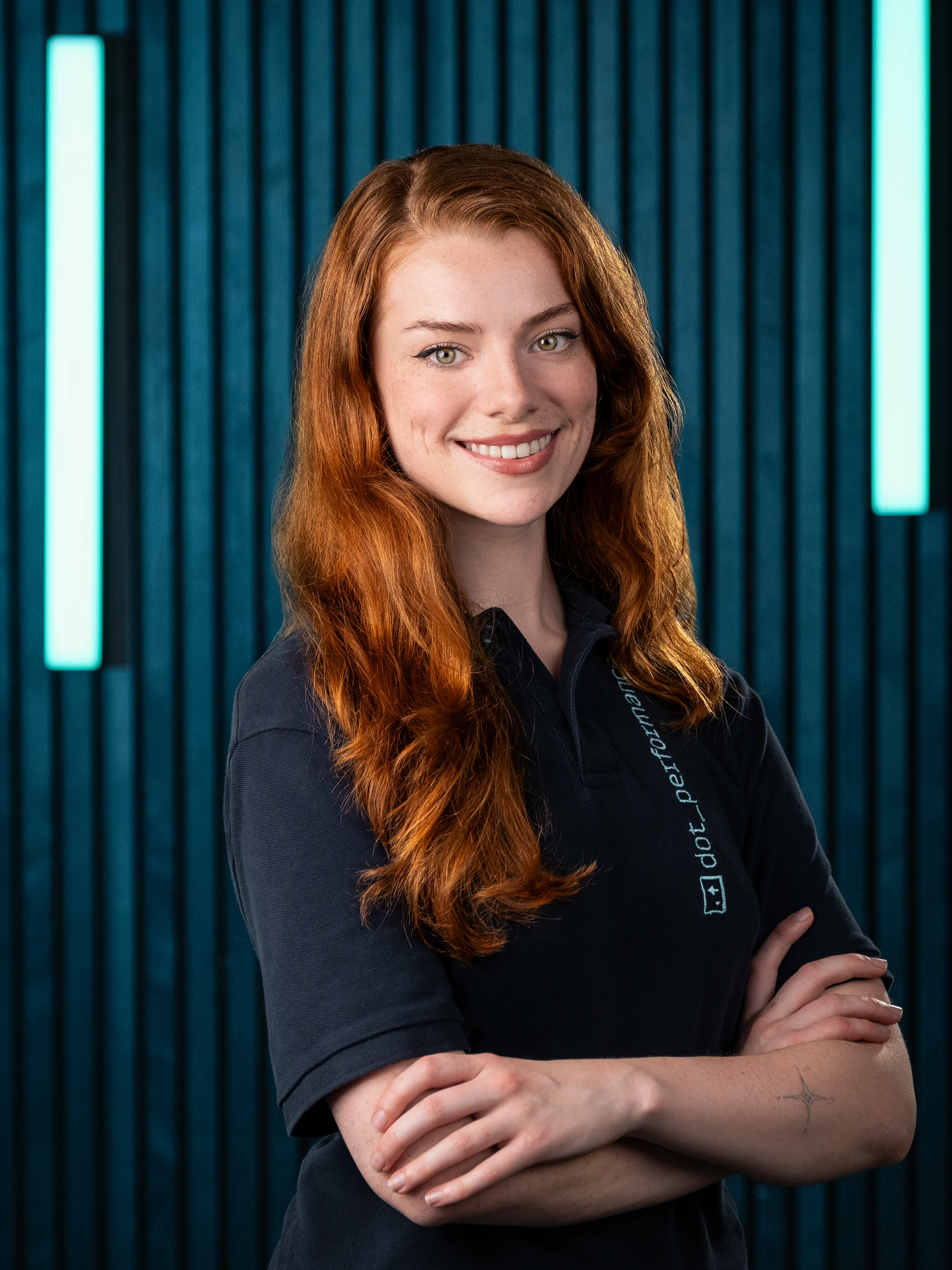 Ella, assistant producer at Dot Performance, smiling with crossed arms as she poses for headshot wearing branded polo shirt.