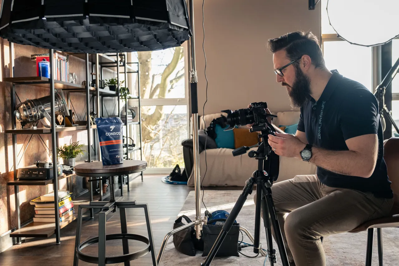 Nico van Loggerenberg photographing a Conrod Coffee product at DotPerformance studios on the Isle of Man, adjusting his camera on a tripod with studio softbox lighting rigged above