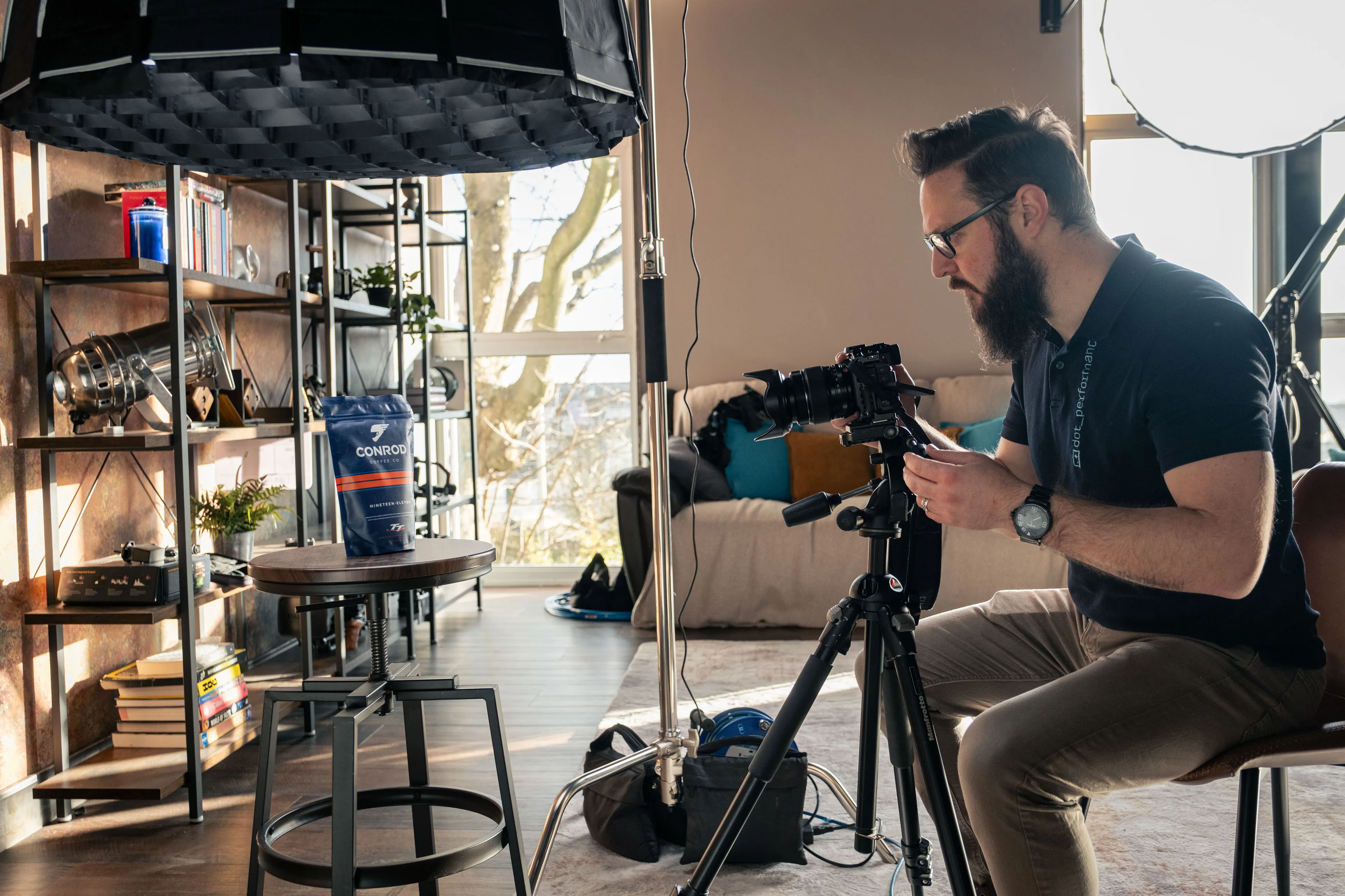 A bearded photographer adjusts a camera on a tripod in an indoor studio setting with lighting equipment and a blue product package displayed on a stool.