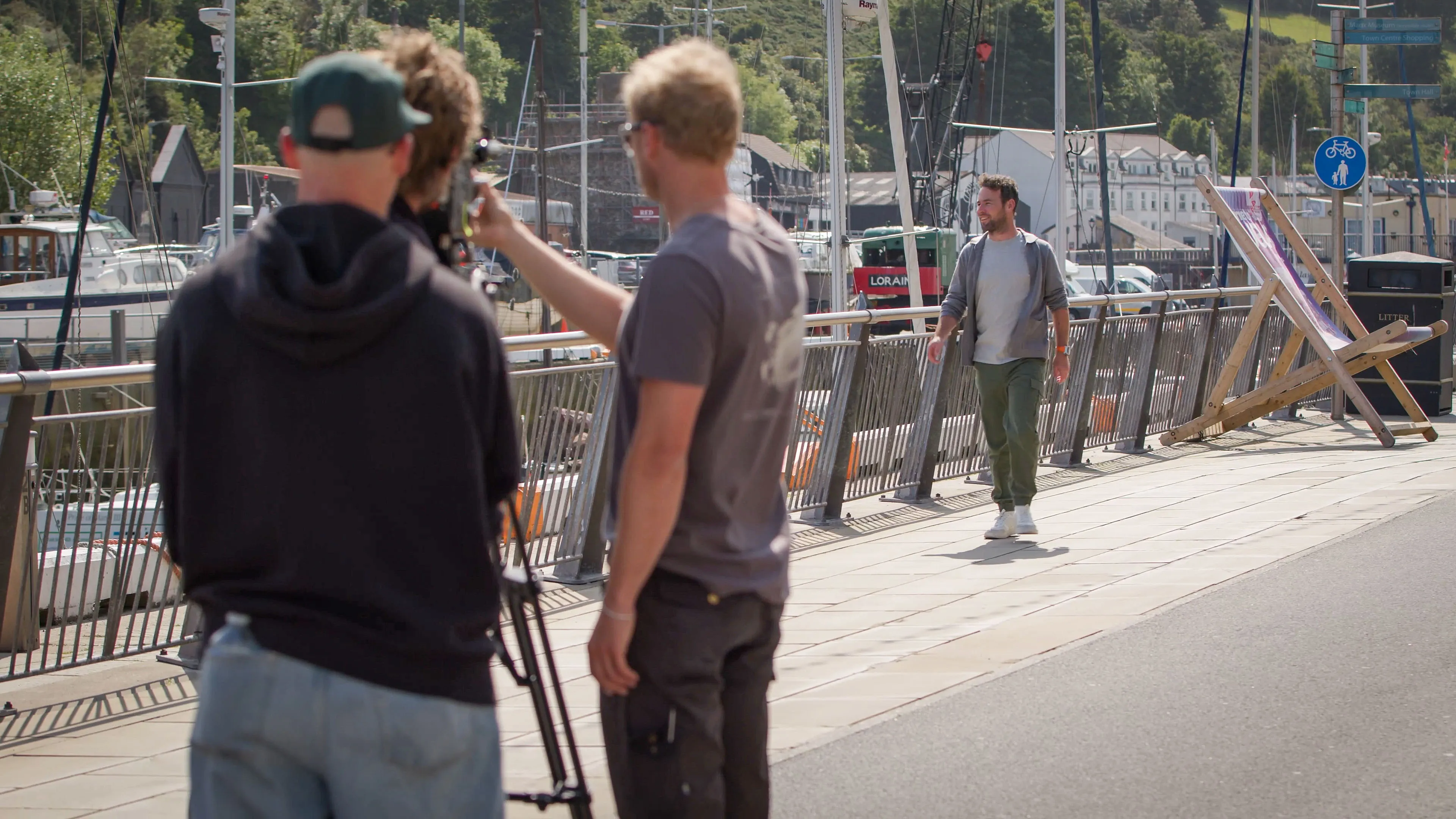 Three men, one with a camera, filming on a sunny marina. A fourth man walks toward them. The scene is calm and professional, set against moored boats and lush hills.