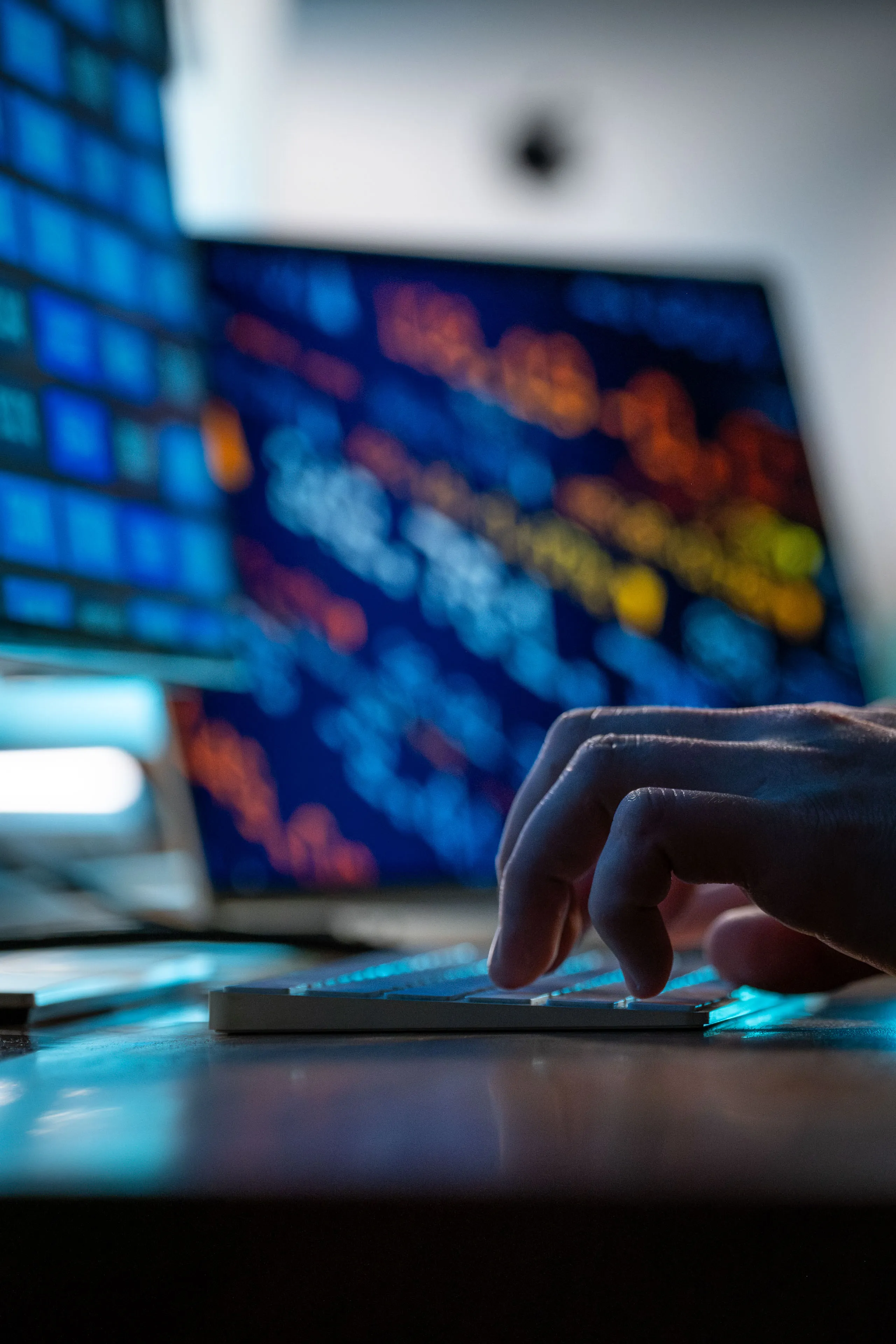 Close-up of hands typing on a laptop keyboard with a blurred, colourful data screen in the background