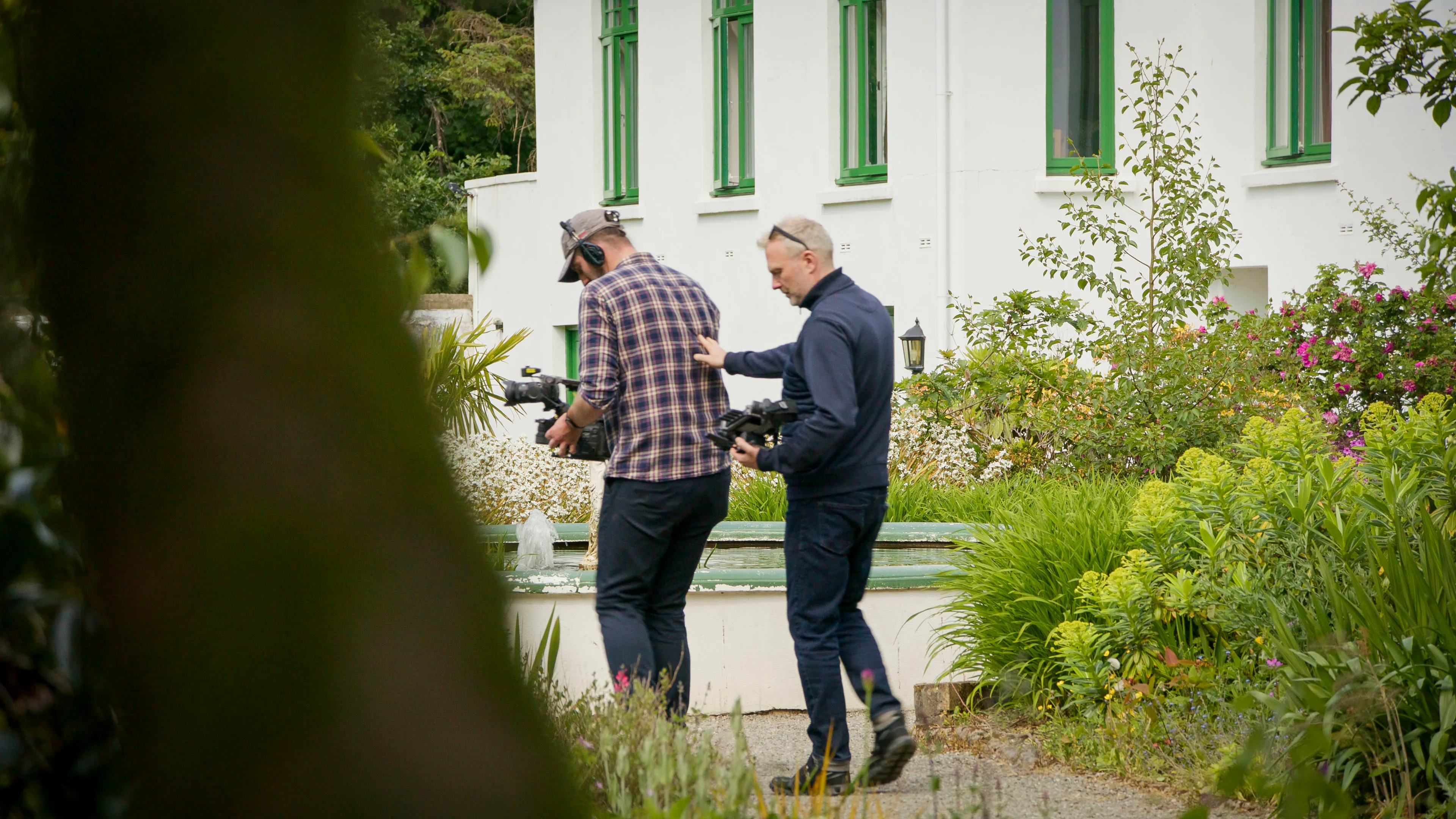 Will Oates walking backwards operating the DJI Ronin 4D camera with the help of the Limehouse director on location in a garden setting on the Isle of Man